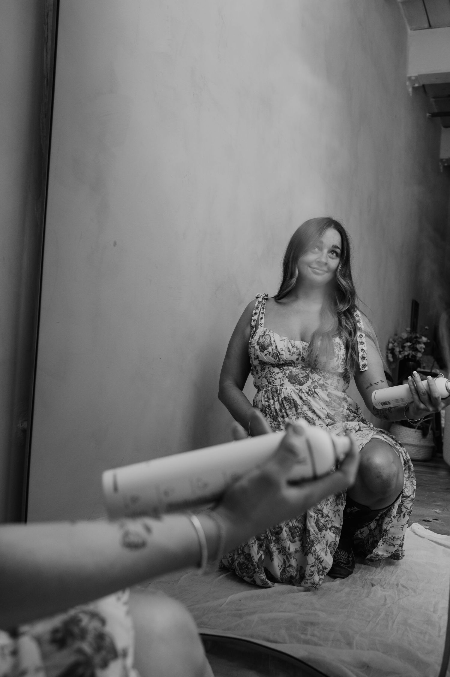 Black and white photo of a smiling woman in a floral dress sitting on the floor as another person extends a cylindrical object toward her.