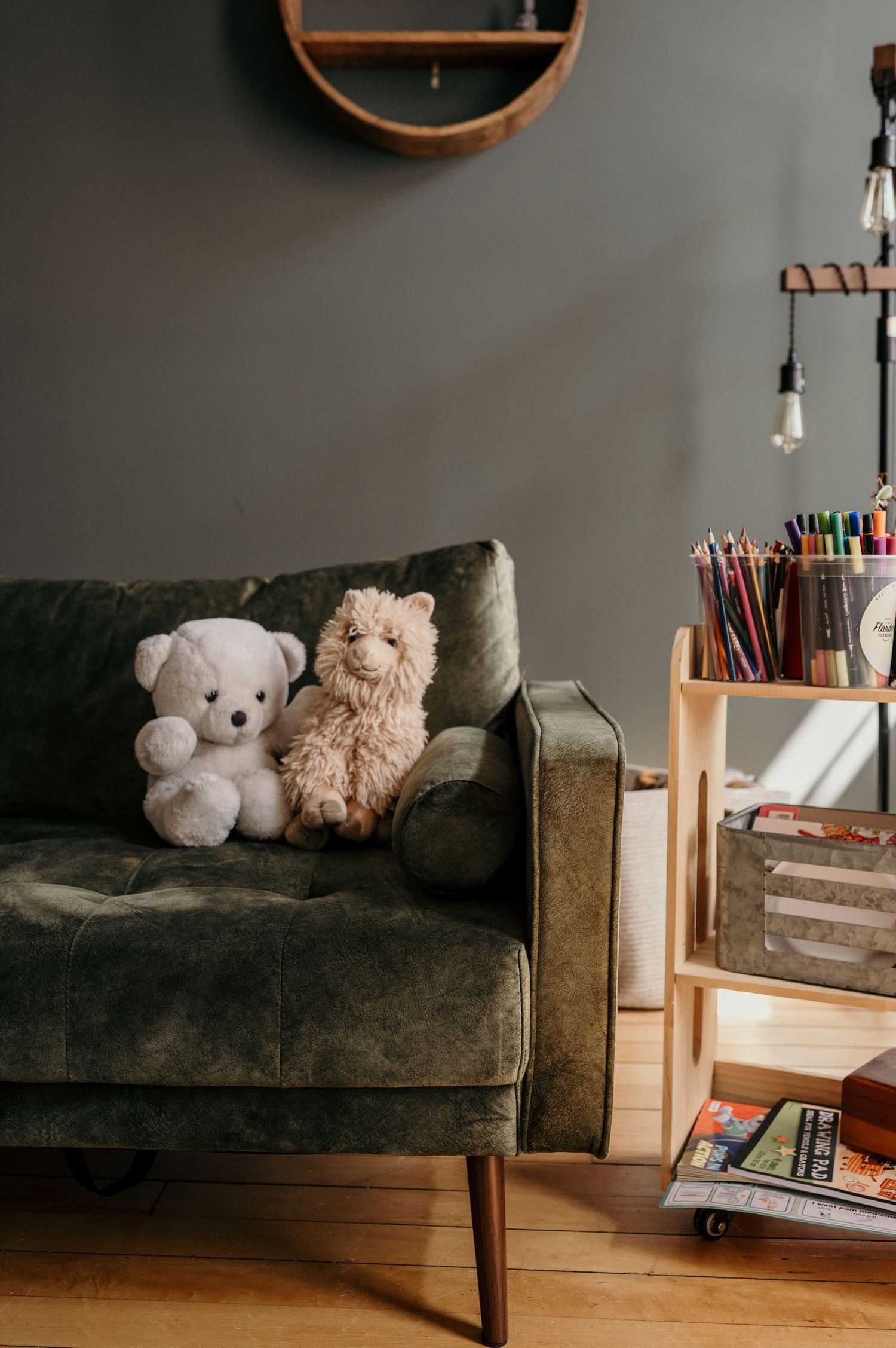 A white teddy bear and a fluffy llama plush sit on a green velvet sofa beside a wooden shelf holding colored pencils and books.