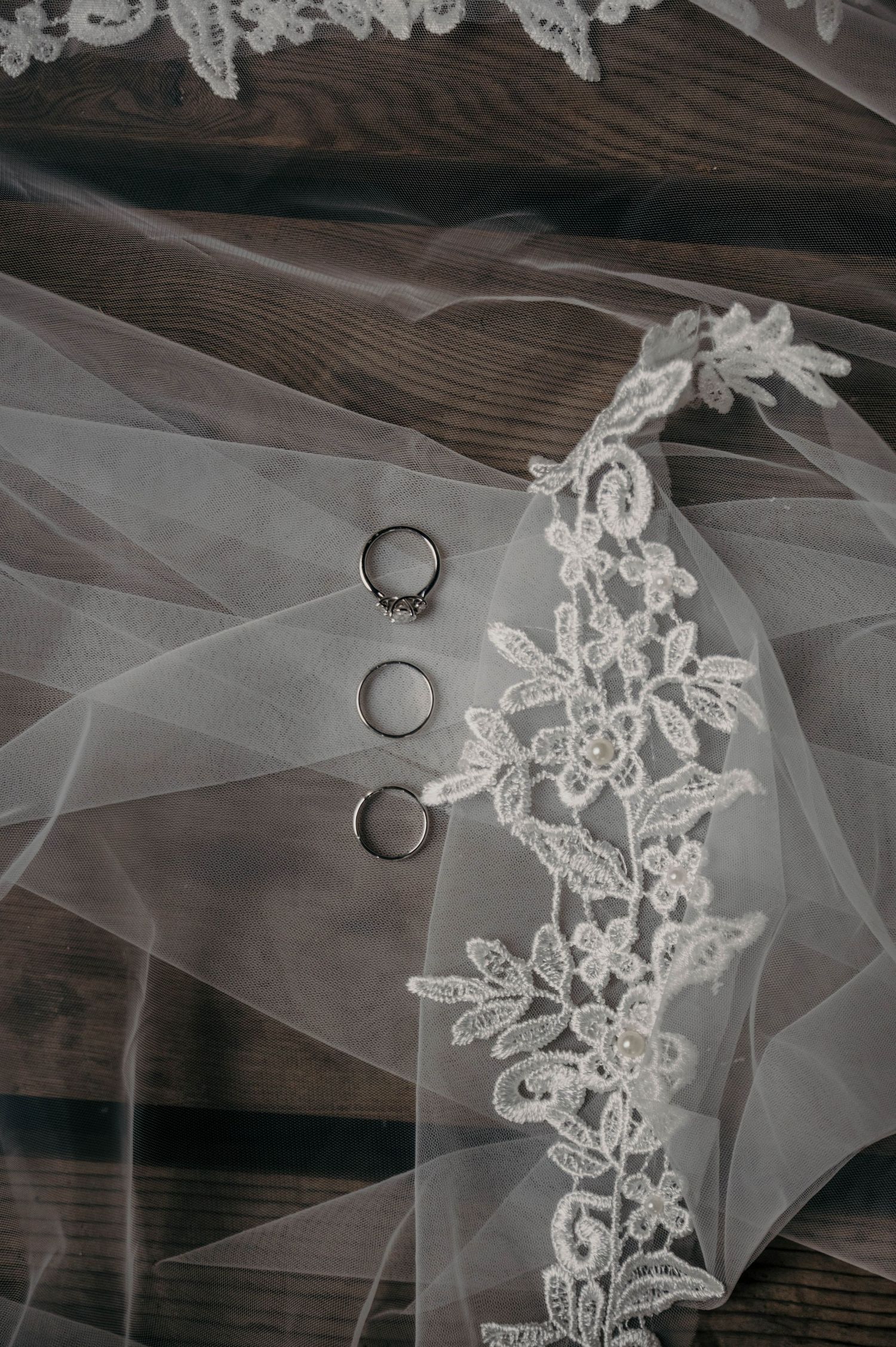 Three wedding rings, including a diamond engagement ring, rest on rustic wood beneath a sheer bridal veil with delicate floral lace trim.