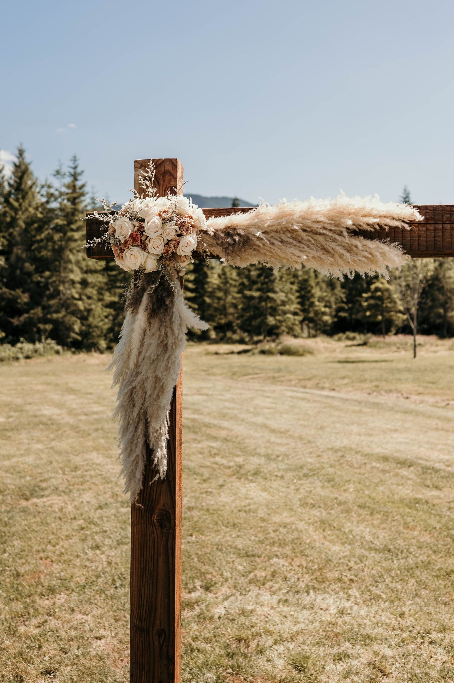Rustic wooden wedding arch corner decorated with pampas grass and cream and blush dried flowers, set in a grassy field with evergreen trees.