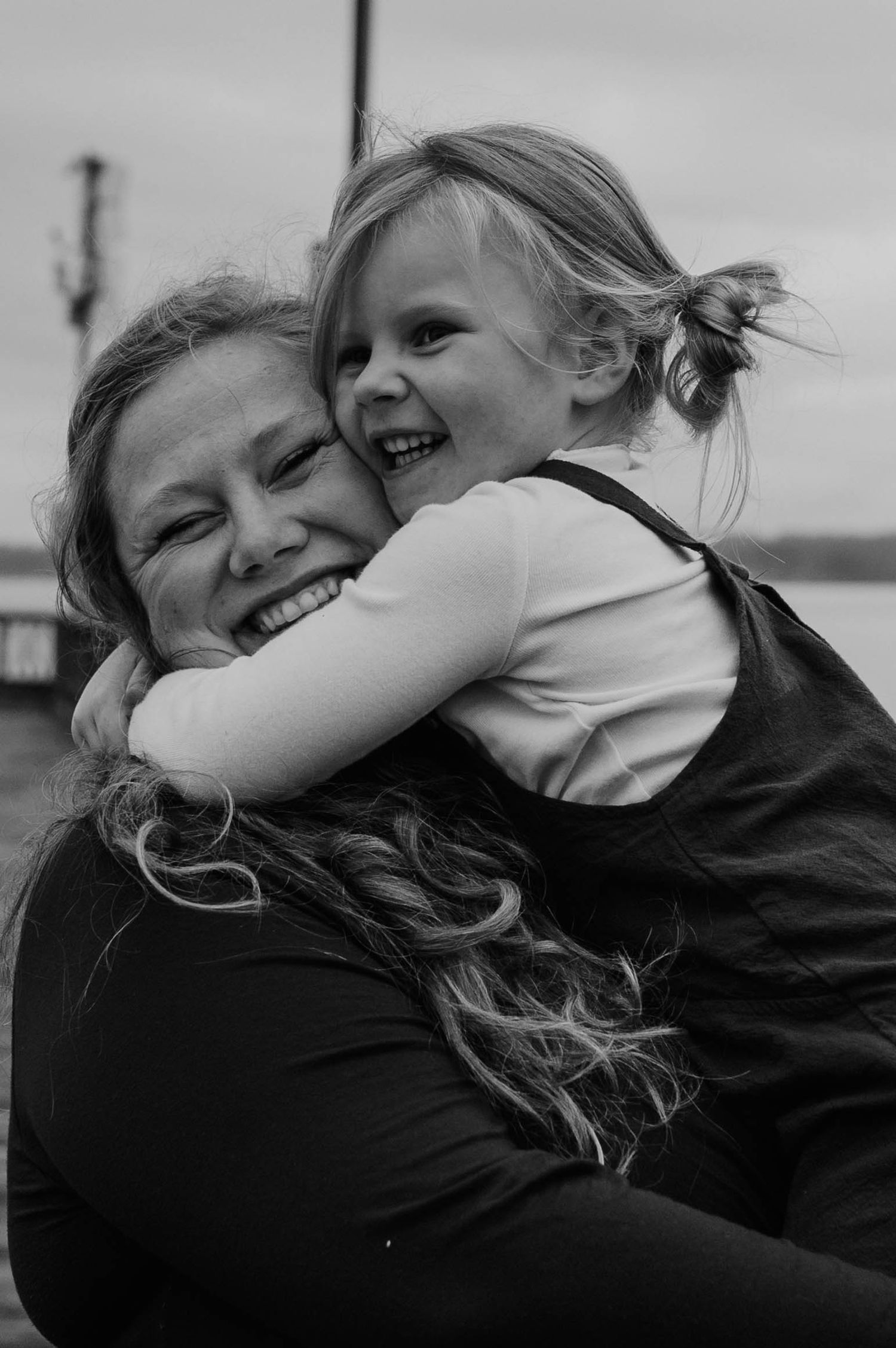 Black and white photo of a smiling woman with long wavy hair hugging a laughing little girl in overalls outdoors near the water.