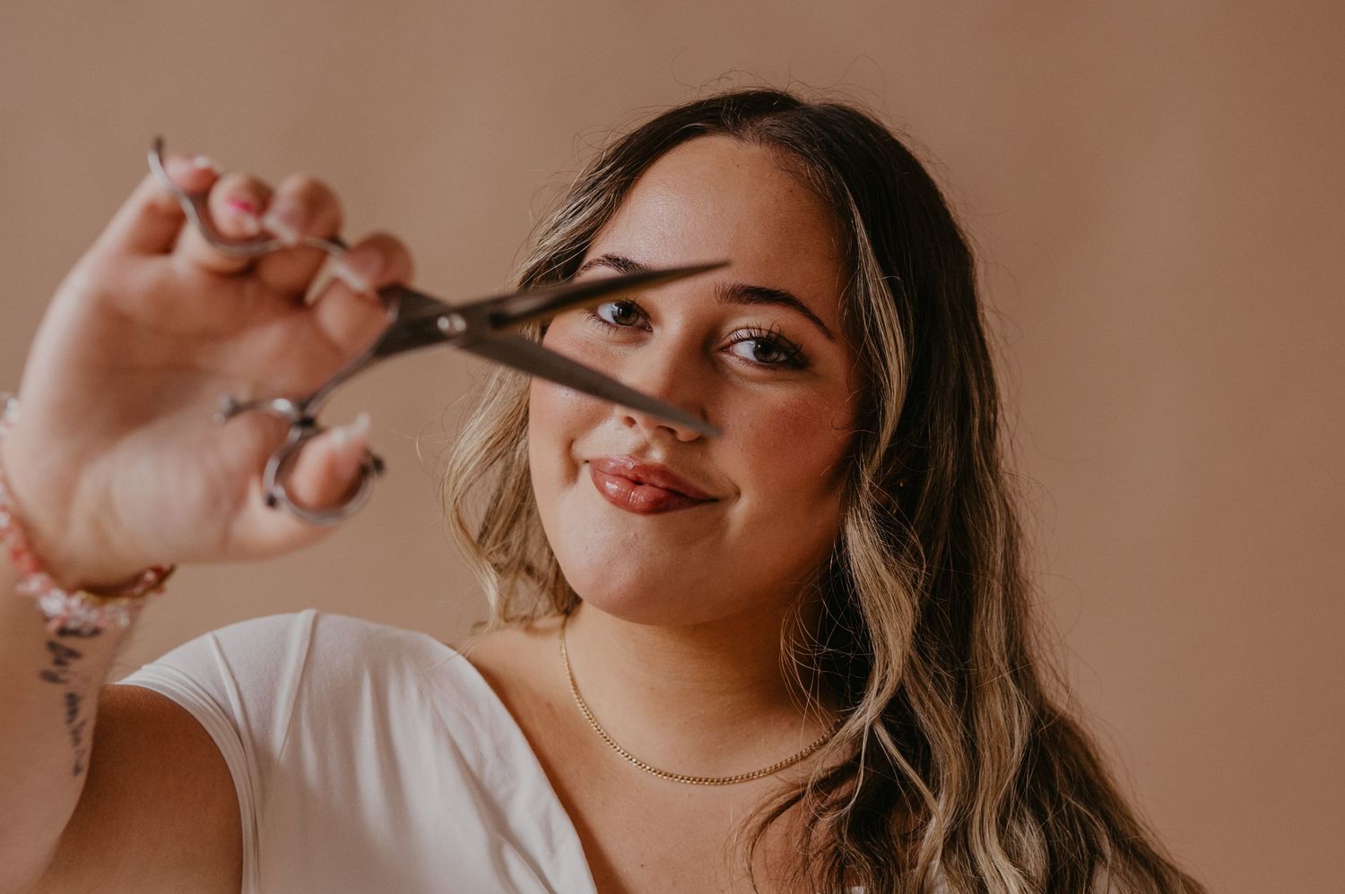 Smiling woman with long wavy hair holds hair cutting shears up to her face, posing against a soft peach background in a white top.
