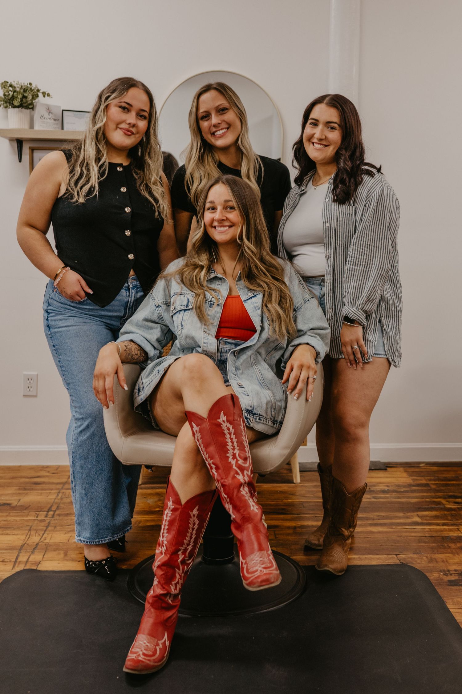 Four smiling women pose in a salon, one seated in a styling chair wearing red cowboy boots and a denim jacket while three stand behind her.