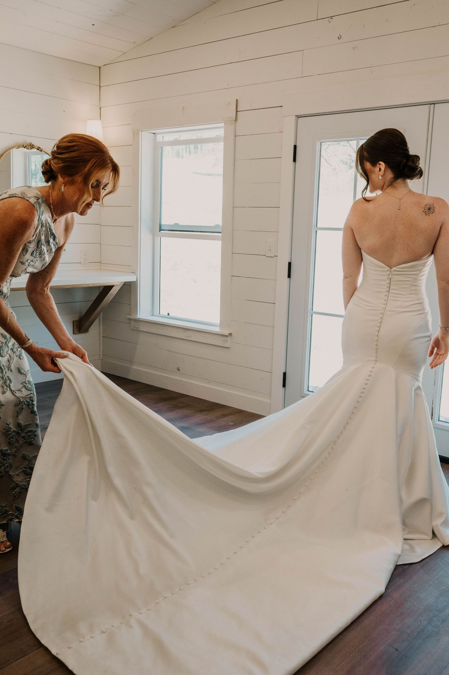 Woman in a floral dress spreads the long train of a bride's strapless white mermaid wedding gown inside a bright rustic room.