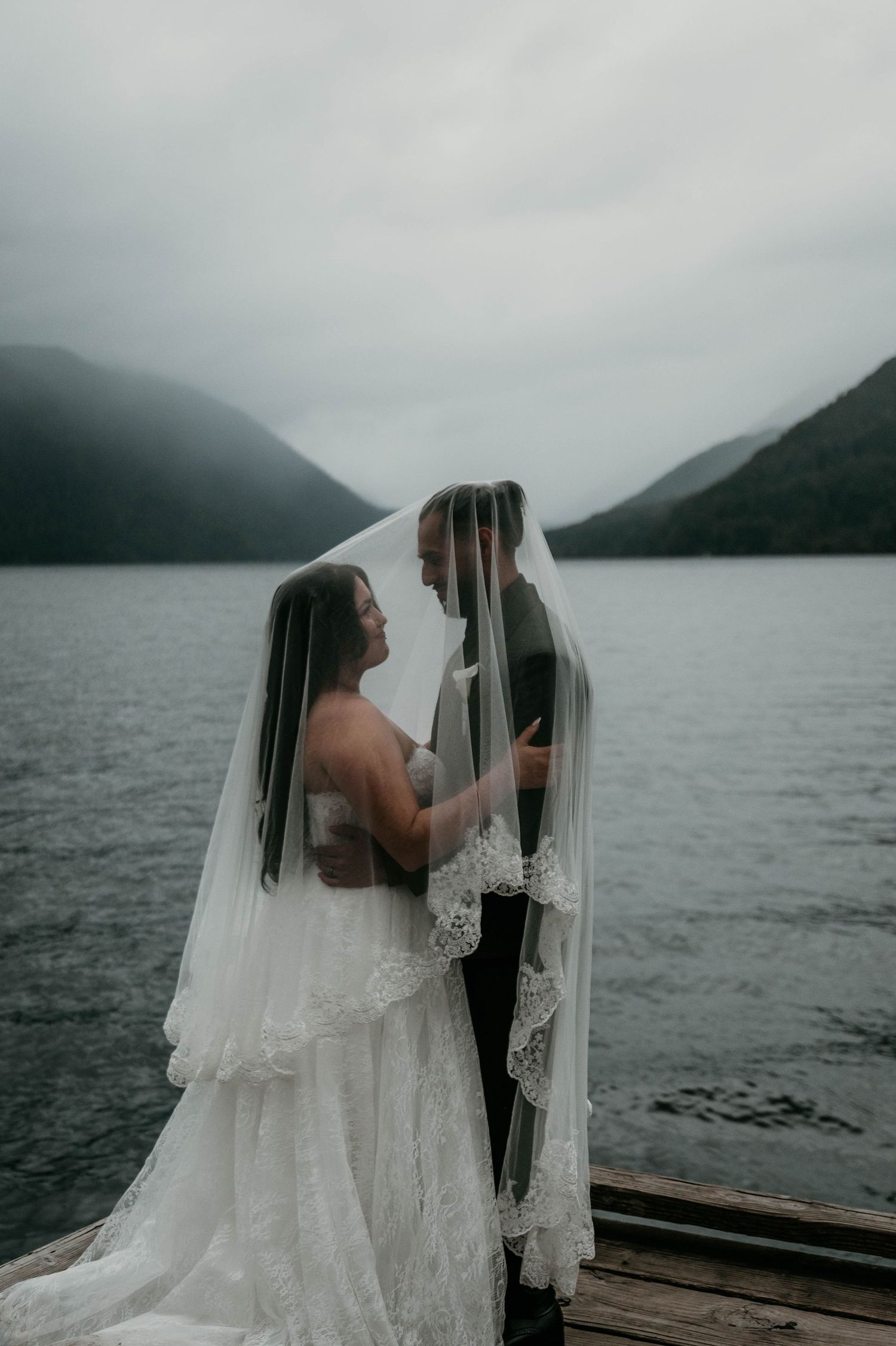 Bride and groom embrace under a lace-trimmed wedding veil on a wooden dock beside a misty mountain lake on a moody overcast day.
