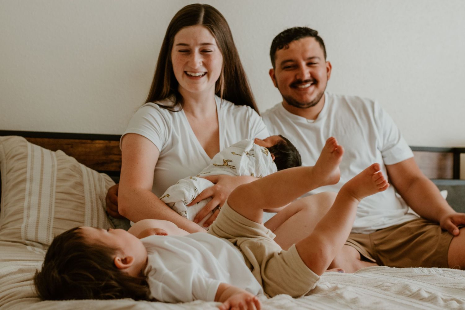 Smiling parents in matching white shirts sit on a bed beside their two young children, who lie playfully with legs kicking in the air.
