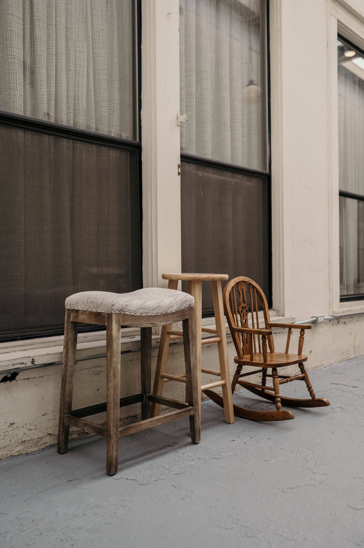 Three vintage wooden seats, a cushioned bar stool, a tall stool and a child's rocking chair, sit on a sidewalk beside a window.