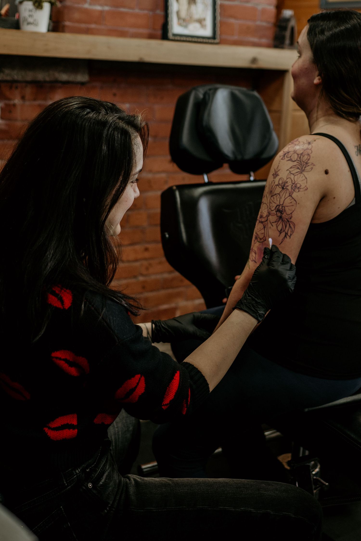 Tattoo artist in black gloves works on a floral tattoo on a client's arm in a cozy studio with brick walls and a black chair.