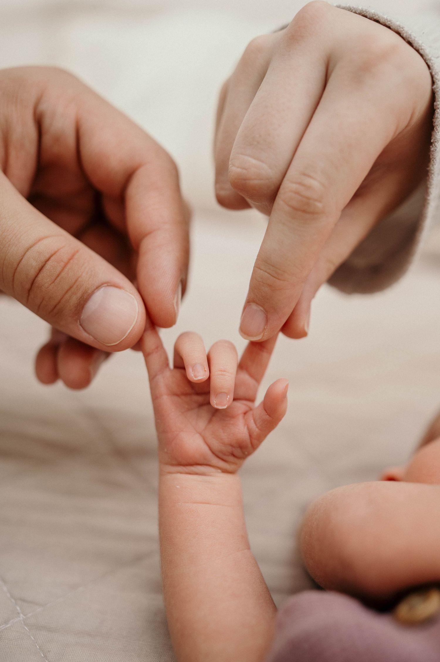 Two adult hands gently touch the tiny fingers of a newborn baby's outstretched hand resting on a soft beige blanket.
