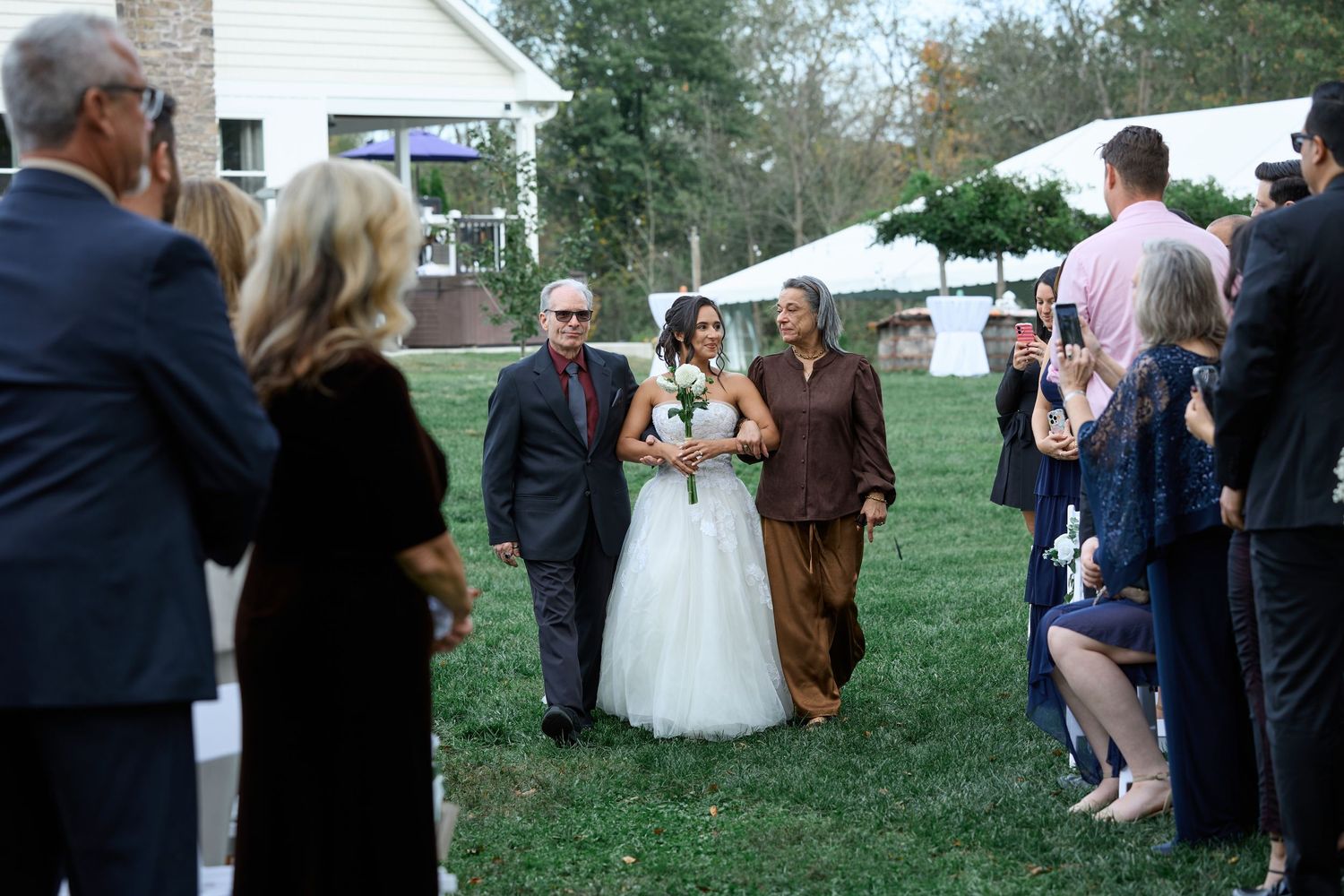 Bride in a white gown walks down the grass aisle between a man in a dark suit and a woman in brown as guests watch.
