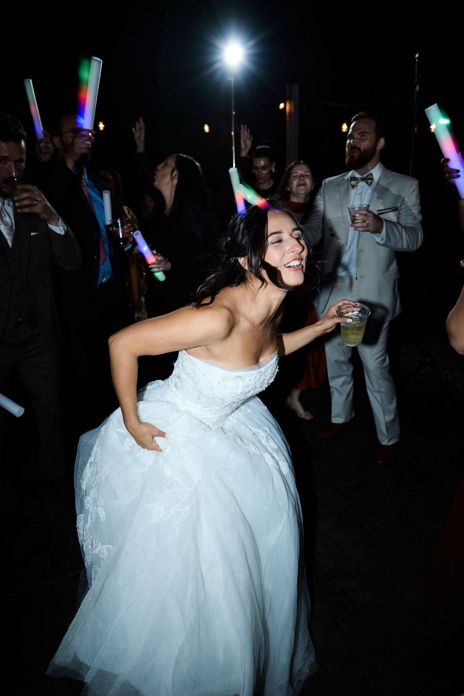 A laughing bride in a strapless white gown dances with a drink in hand as guests wave glow sticks behind her at night.