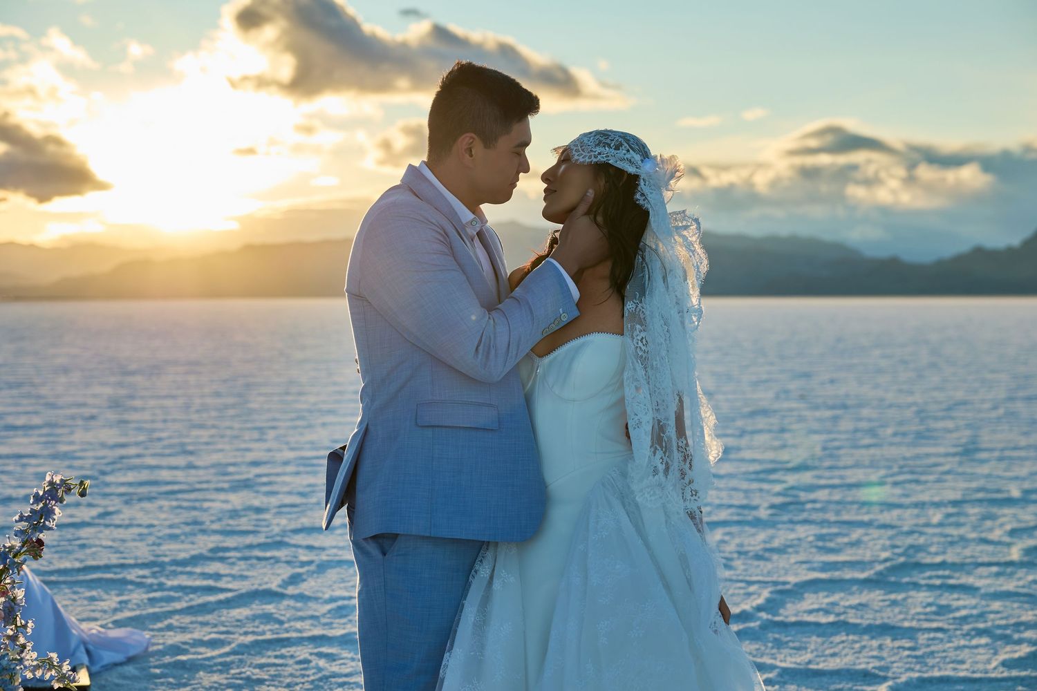 Groom in a blue suit leans in to kiss his veiled bride as the sun sets over a glassy salt flat.