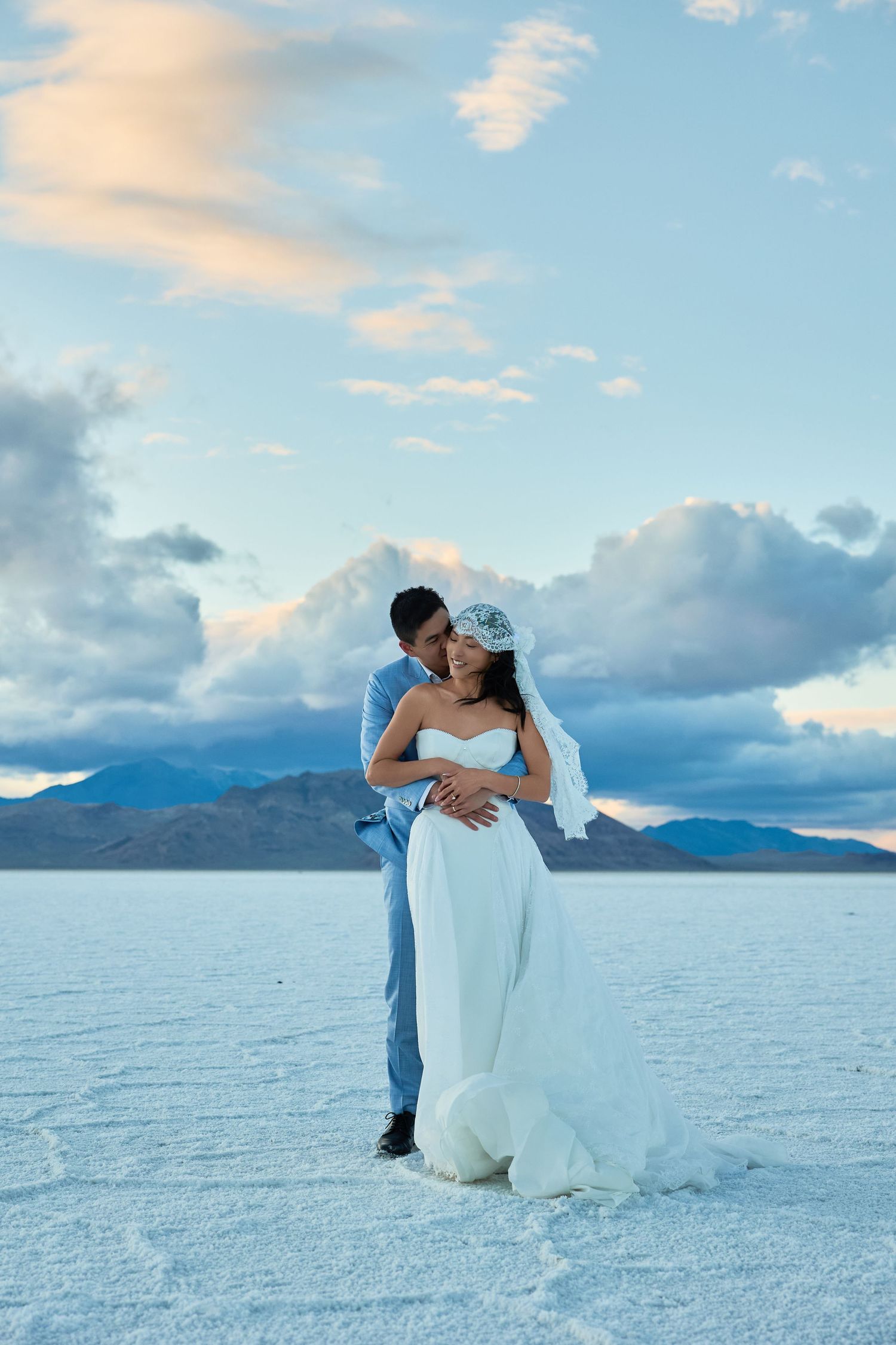Groom in a blue suit embraces the bride in a flowing white wedding dress on a vast white salt flat with mountains behind.
