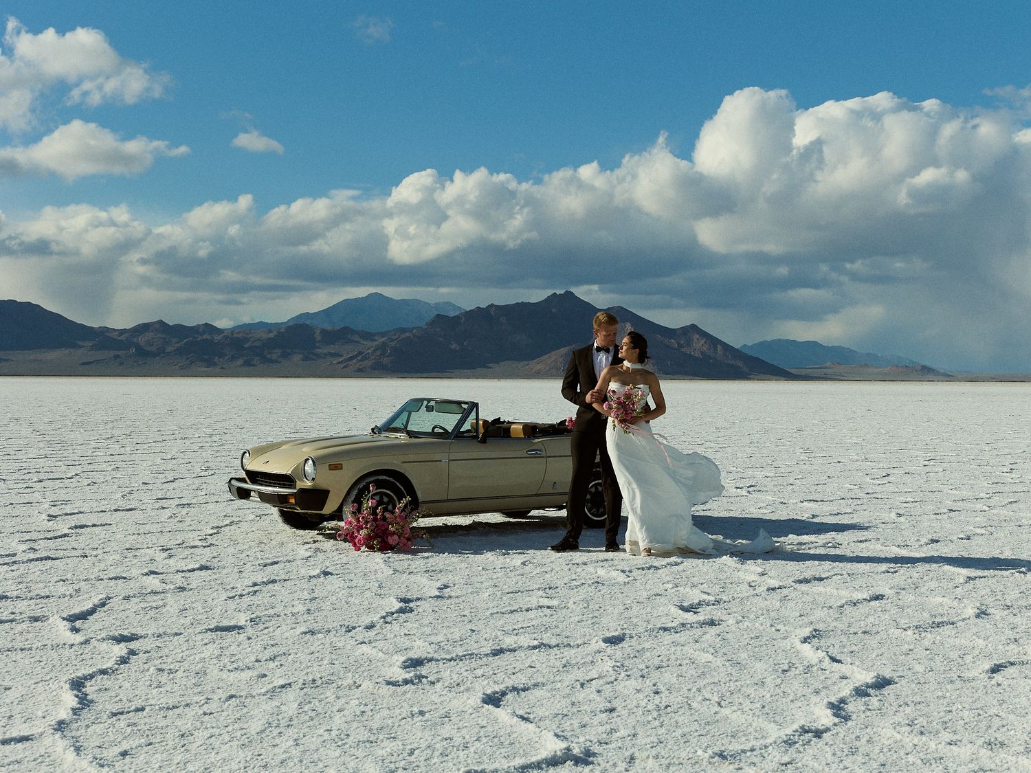 A bride in a white gown and groom in a tuxedo stand by a vintage convertible on a salt flat with dramatic mountains behind.