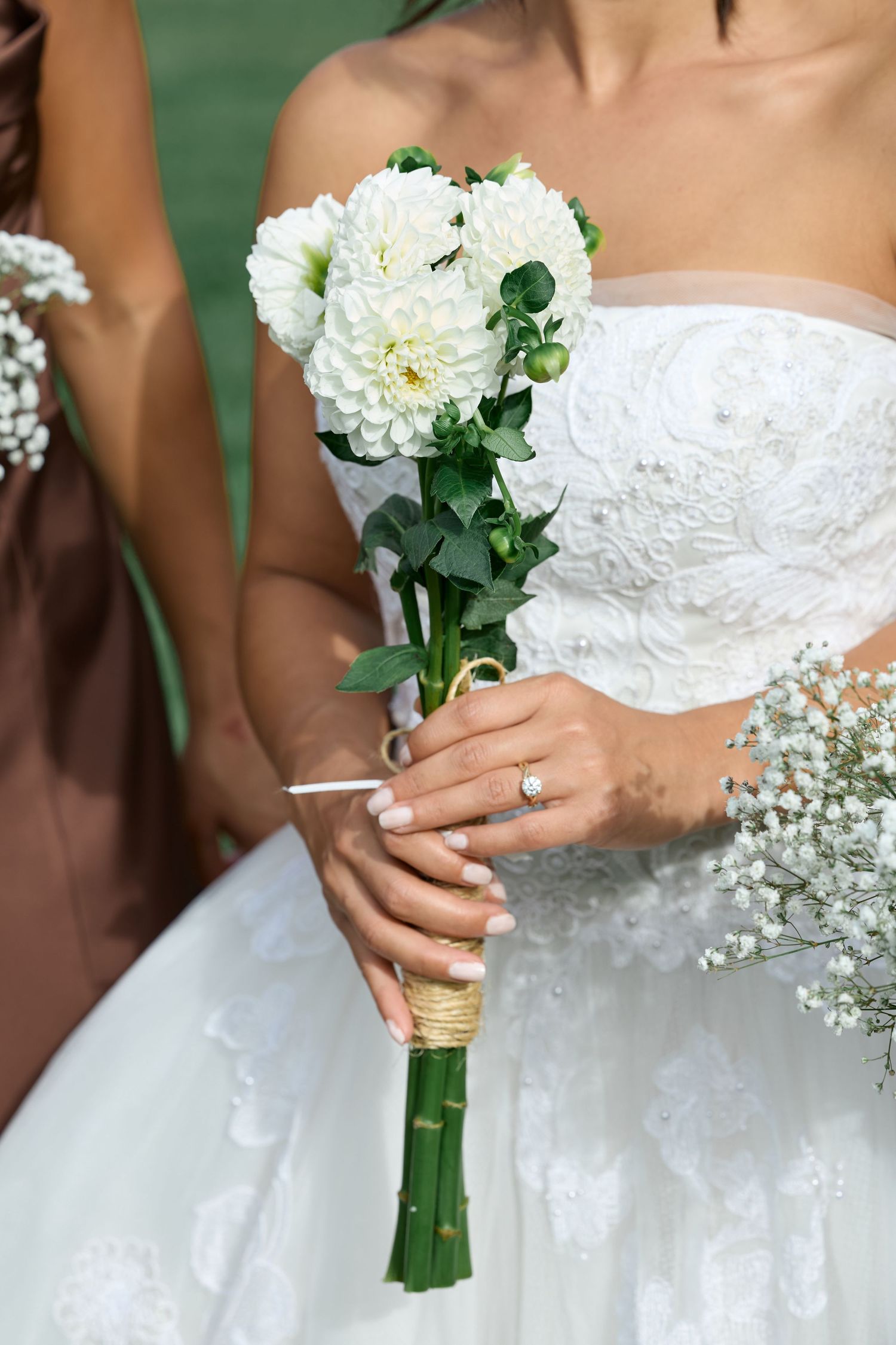 Close-up of a bride's hands with a diamond ring holding a white dahlia bouquet wrapped in twine in front of her lace gown.