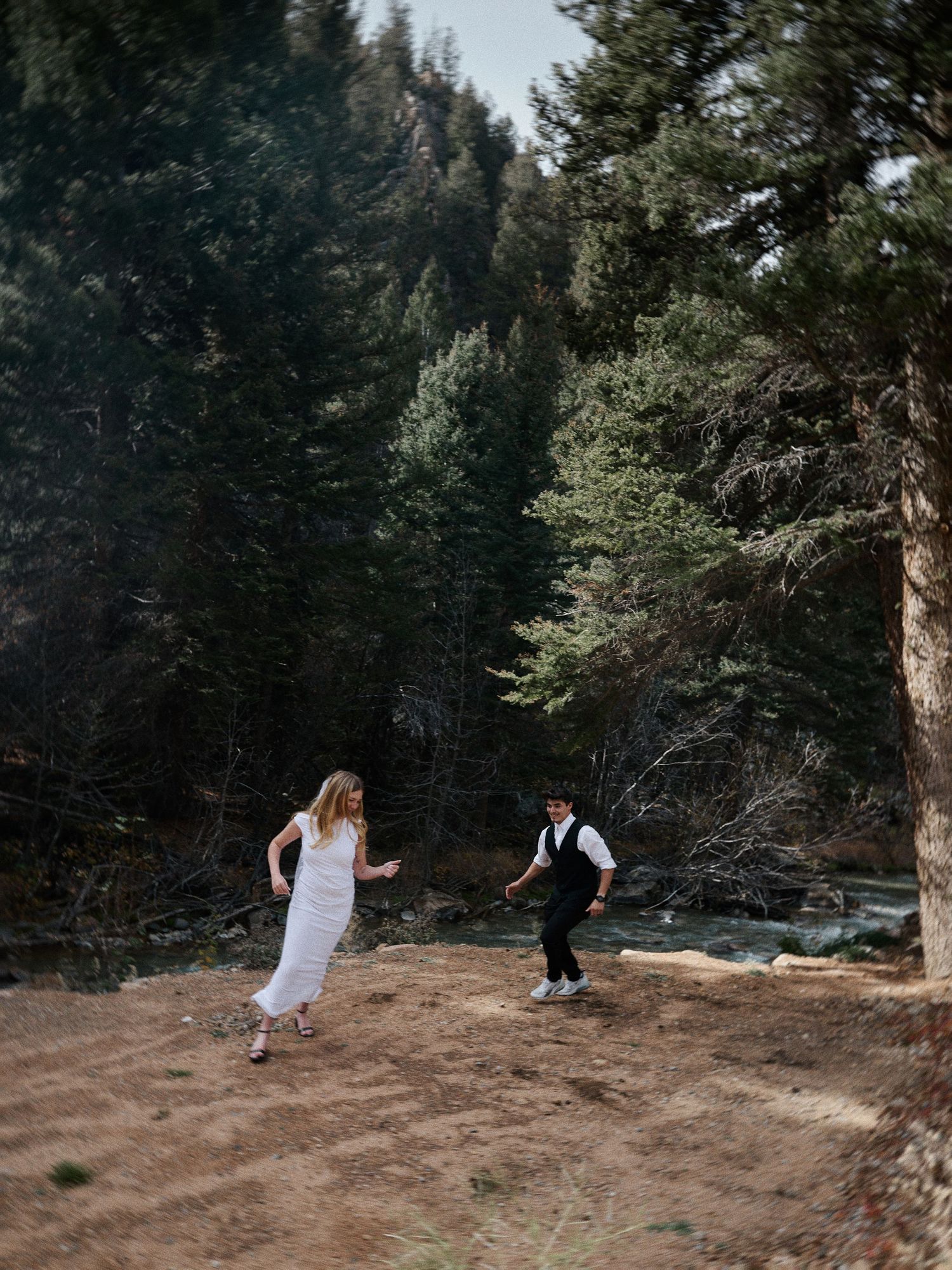 Bride in a white dress and groom in a vest walk playfully along a forest clearing beside a stream during their elopement.