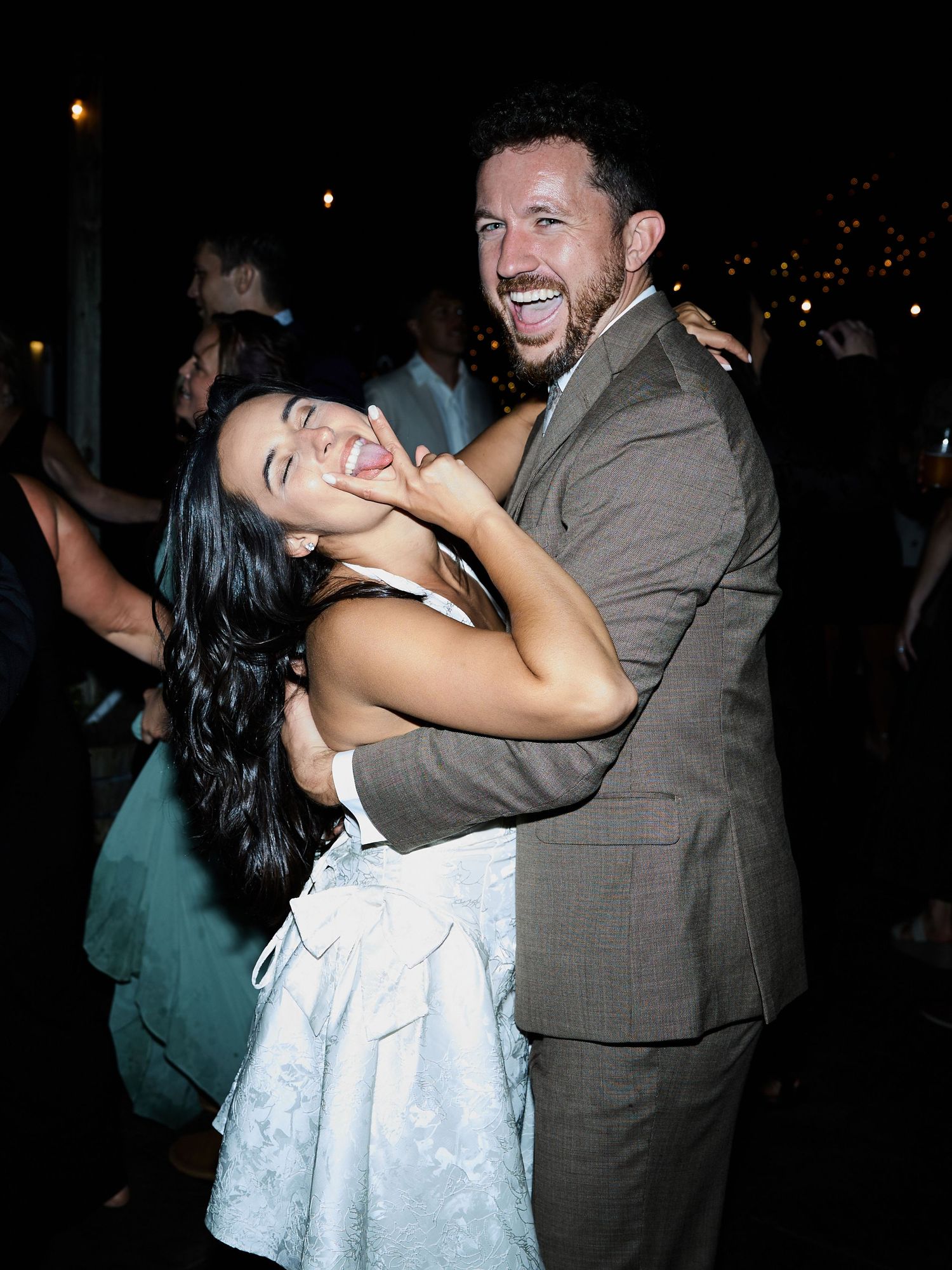 A laughing couple embraces on the dance floor, the woman in a white dress and the man in a brown suit.