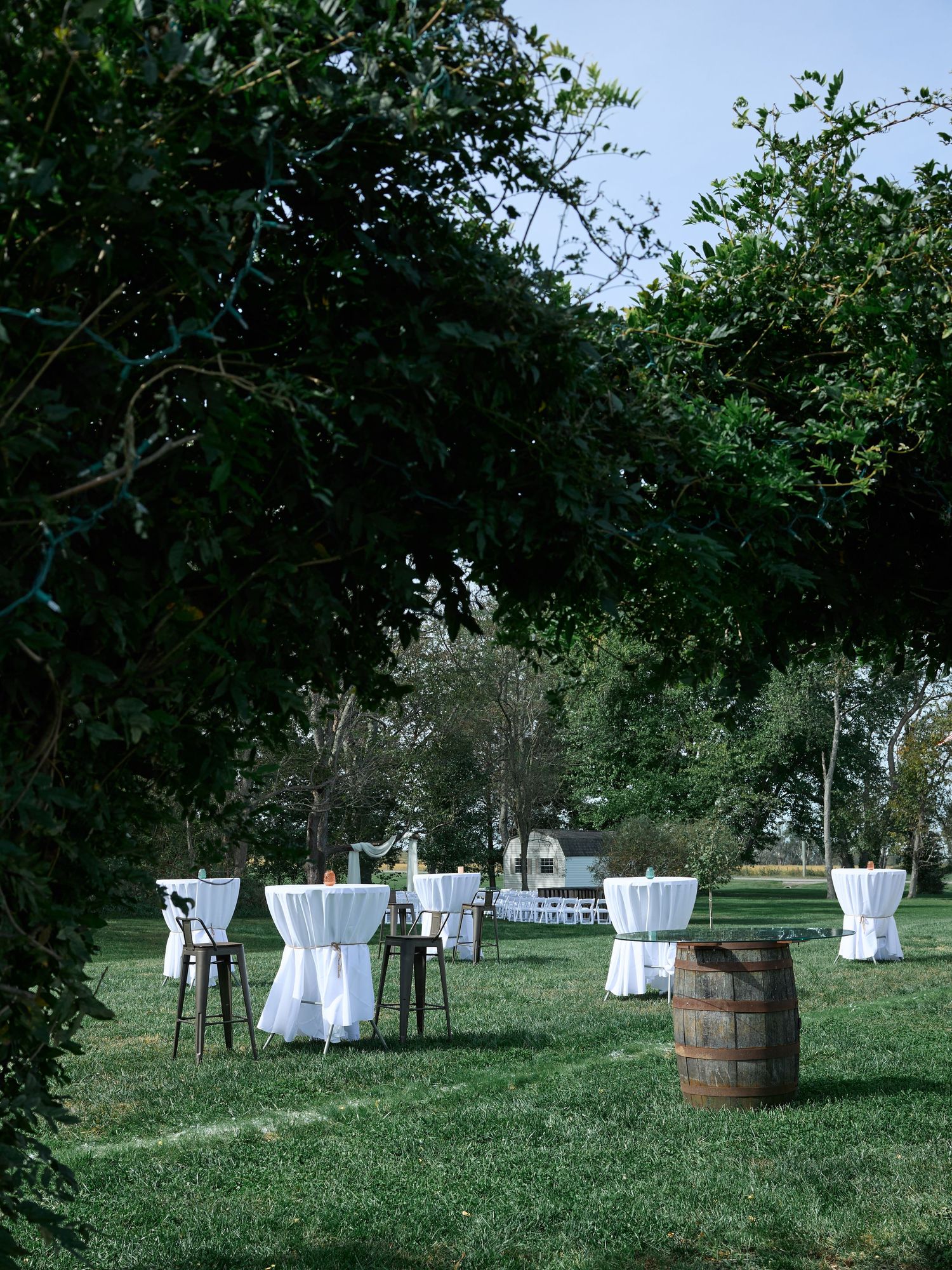 White-clothed cocktail tables, bar stools, and a wooden barrel on a lawn under leafy branches at an outdoor wedding reception.