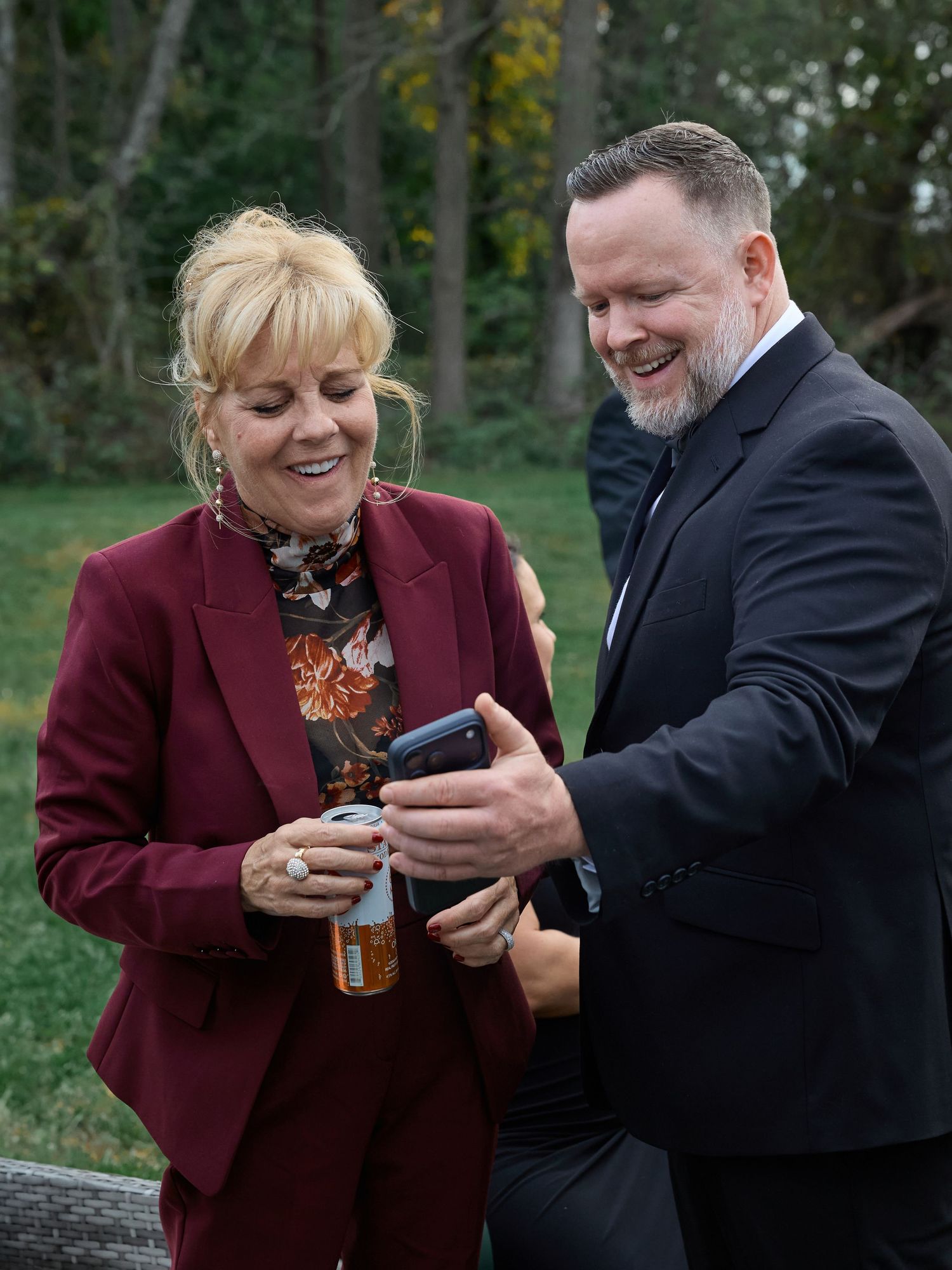 A smiling woman in a burgundy pantsuit holding a drink looks at a phone shown by a bearded man in a dark suit outdoors.