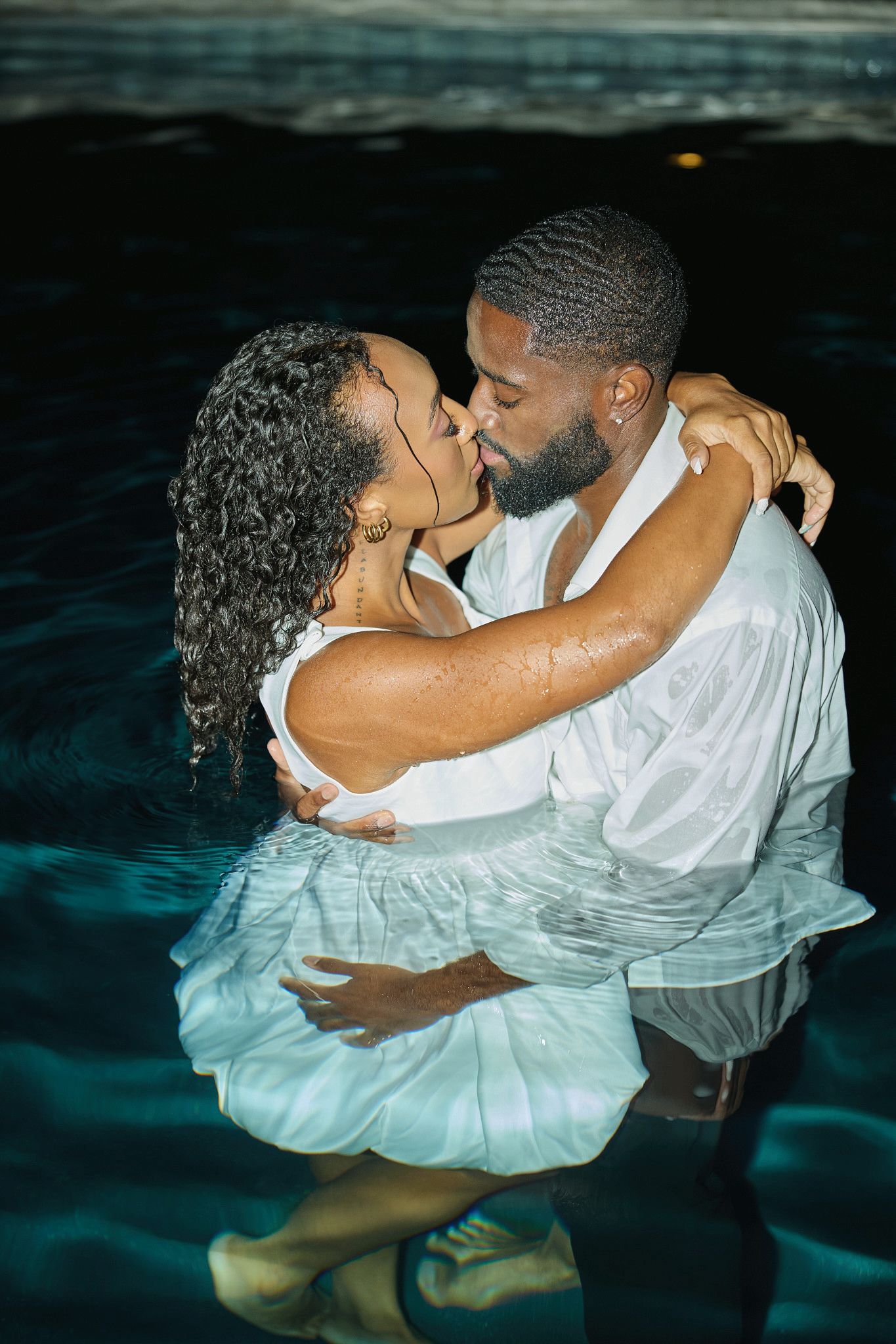 A couple in wet white outfits shares a kiss while standing waist-deep in a dark nighttime pool.
