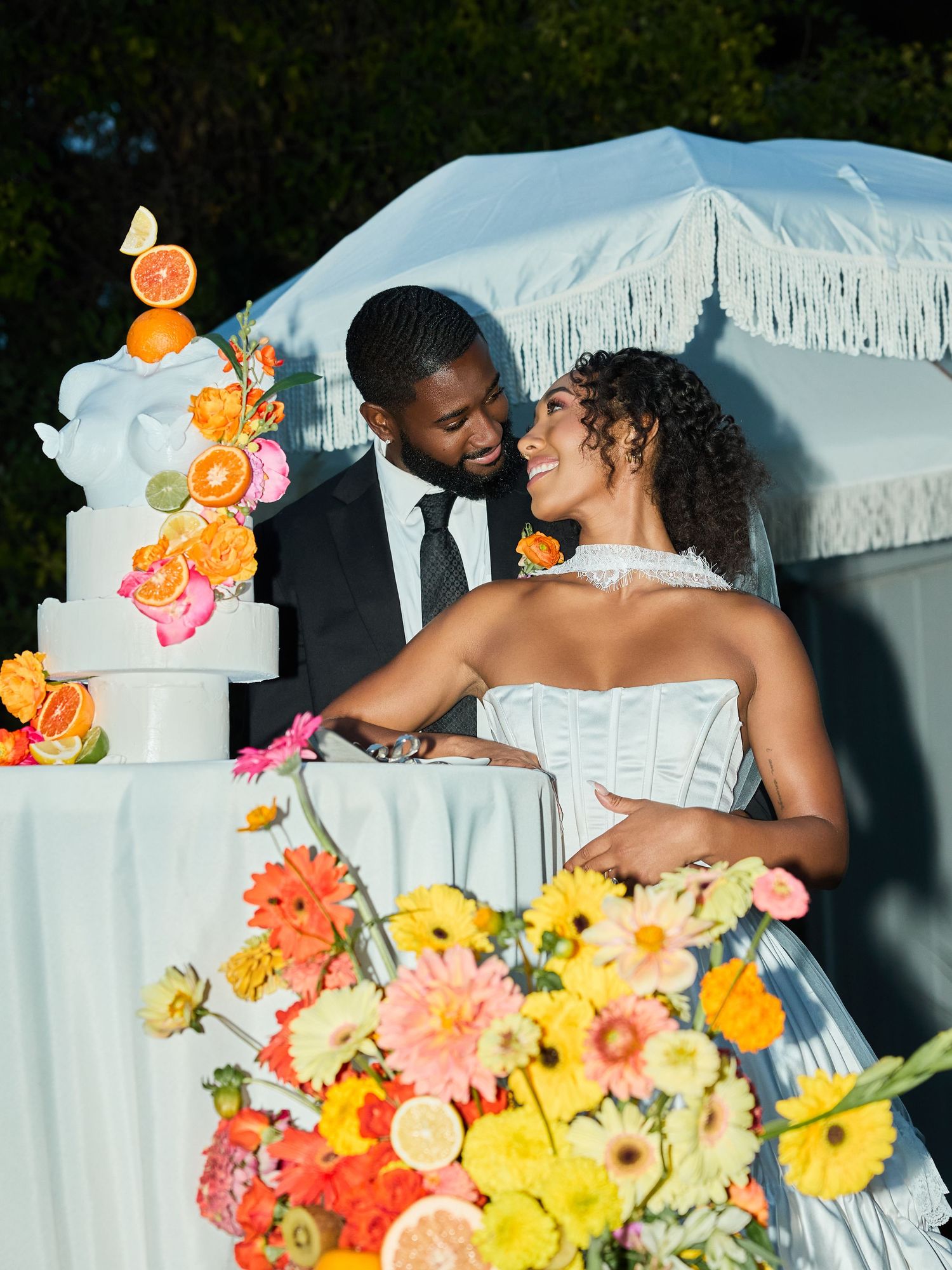 Bride and groom share a joyful moment beside a fringed white umbrella, a citrus-decorated wedding cake, and colorful gerbera daisies.