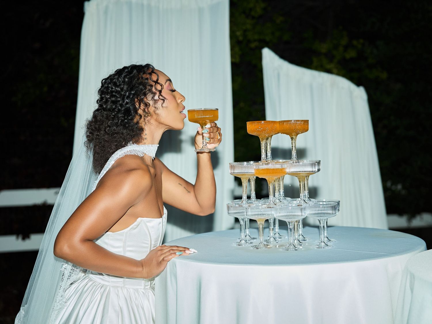 A bride in a white wedding dress sips from a champagne coupe beside a glass tower at a draped table during an outdoor evening reception.