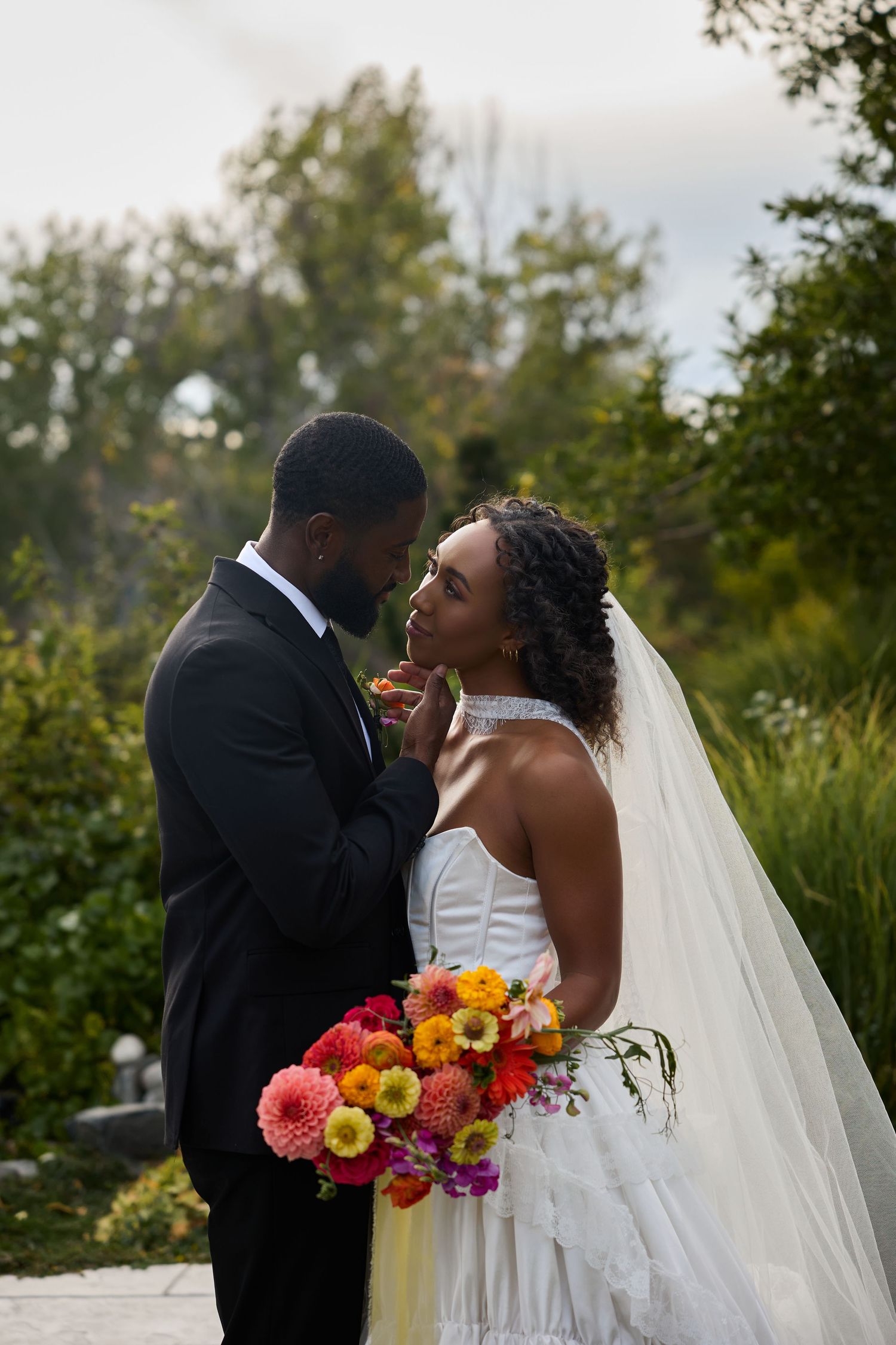 Groom in a black suit tenderly touches the bride's face as she holds a bright floral bouquet in a garden.