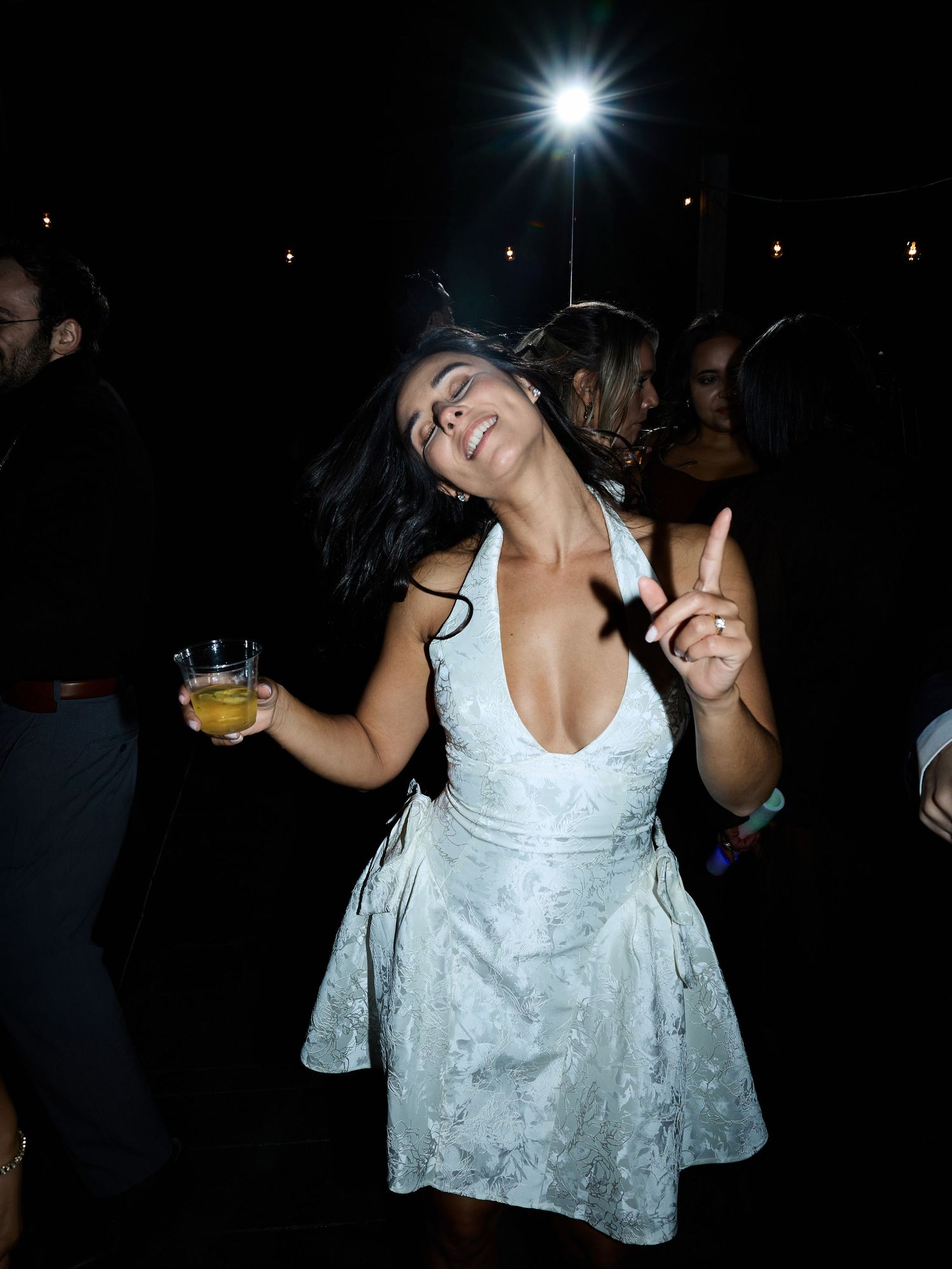 A woman in a white floral mini dress dances joyfully with a drink in hand at a night wedding reception.