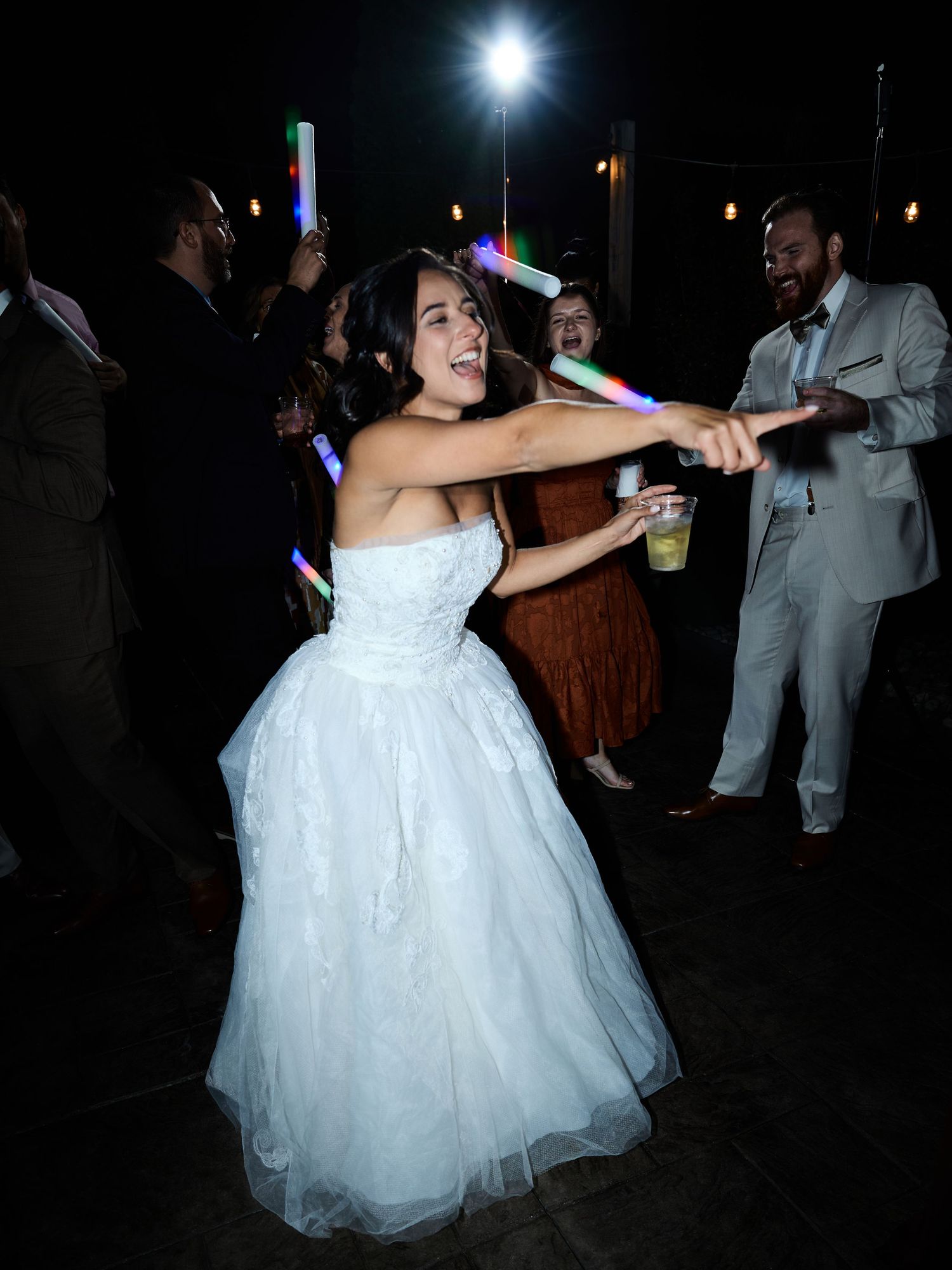A smiling bride in a white lace gown points while dancing, holding a drink as guests with glow sticks celebrate nearby.