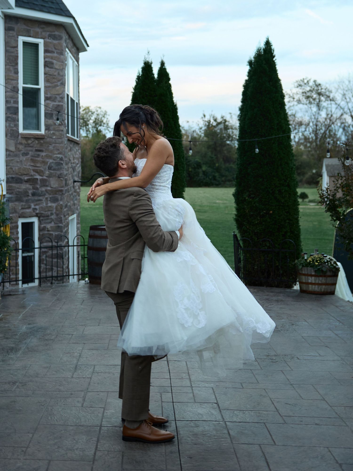 A groom in a brown suit lifts his smiling bride in a flowing white wedding dress on a stone patio beside a manor house.