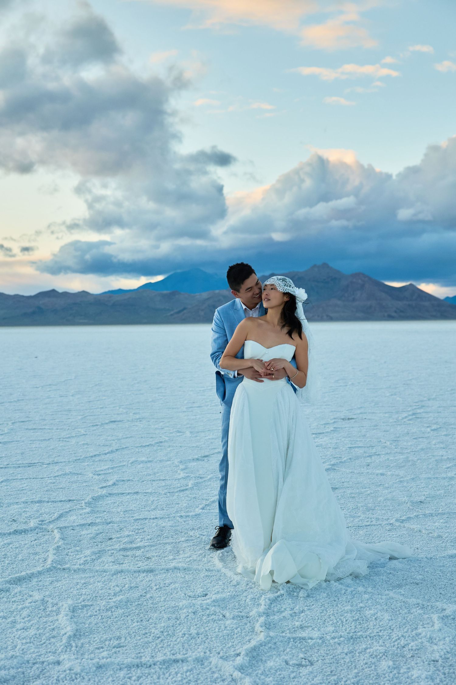 Groom holds the bride from behind as she wears a white gown and veil, standing on cracked white salt flats beneath dramatic clouds.