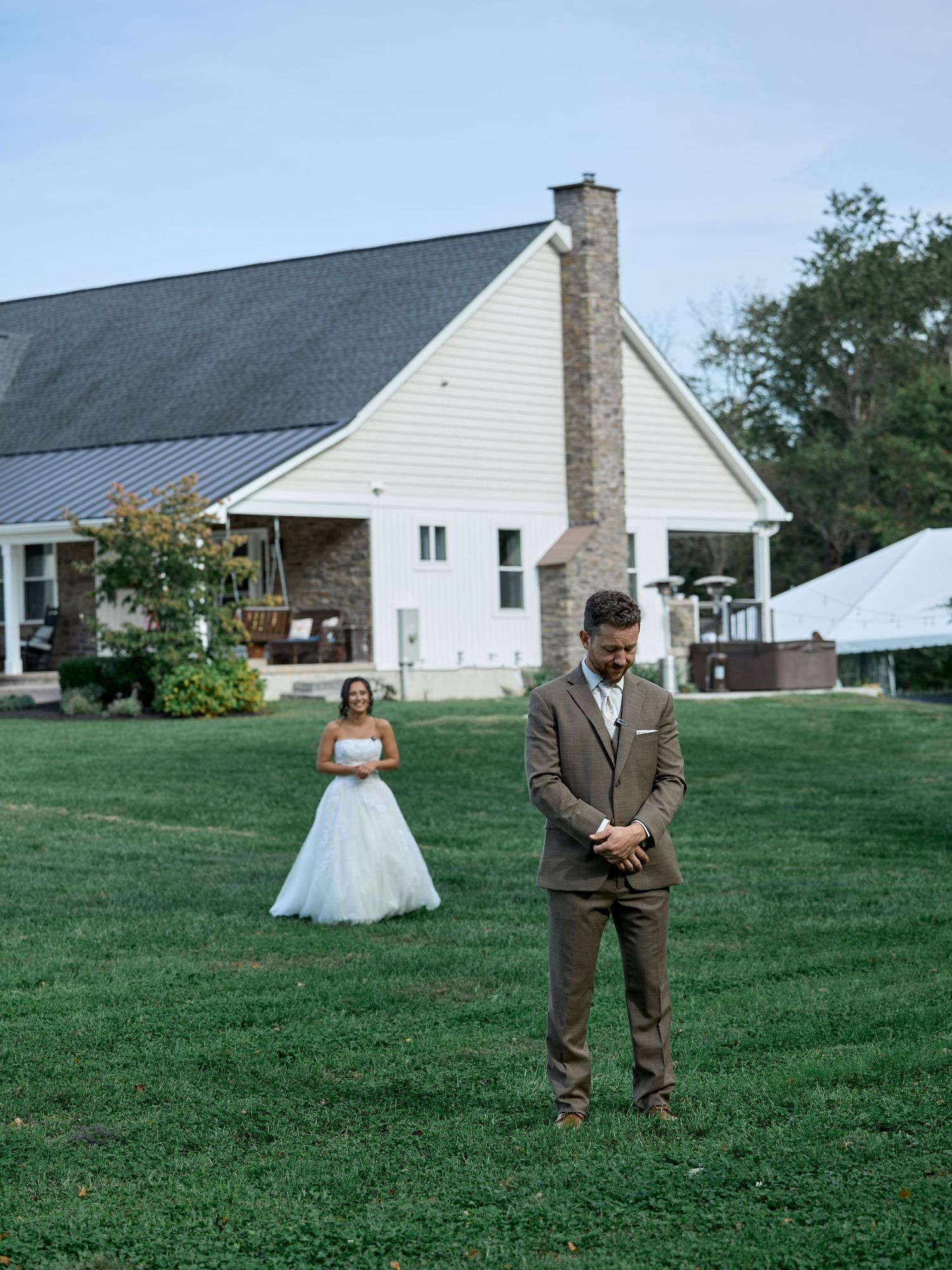 Groom in a brown suit faces away with his head bowed while the bride in a white gown waits behind him in front of a farmhouse.