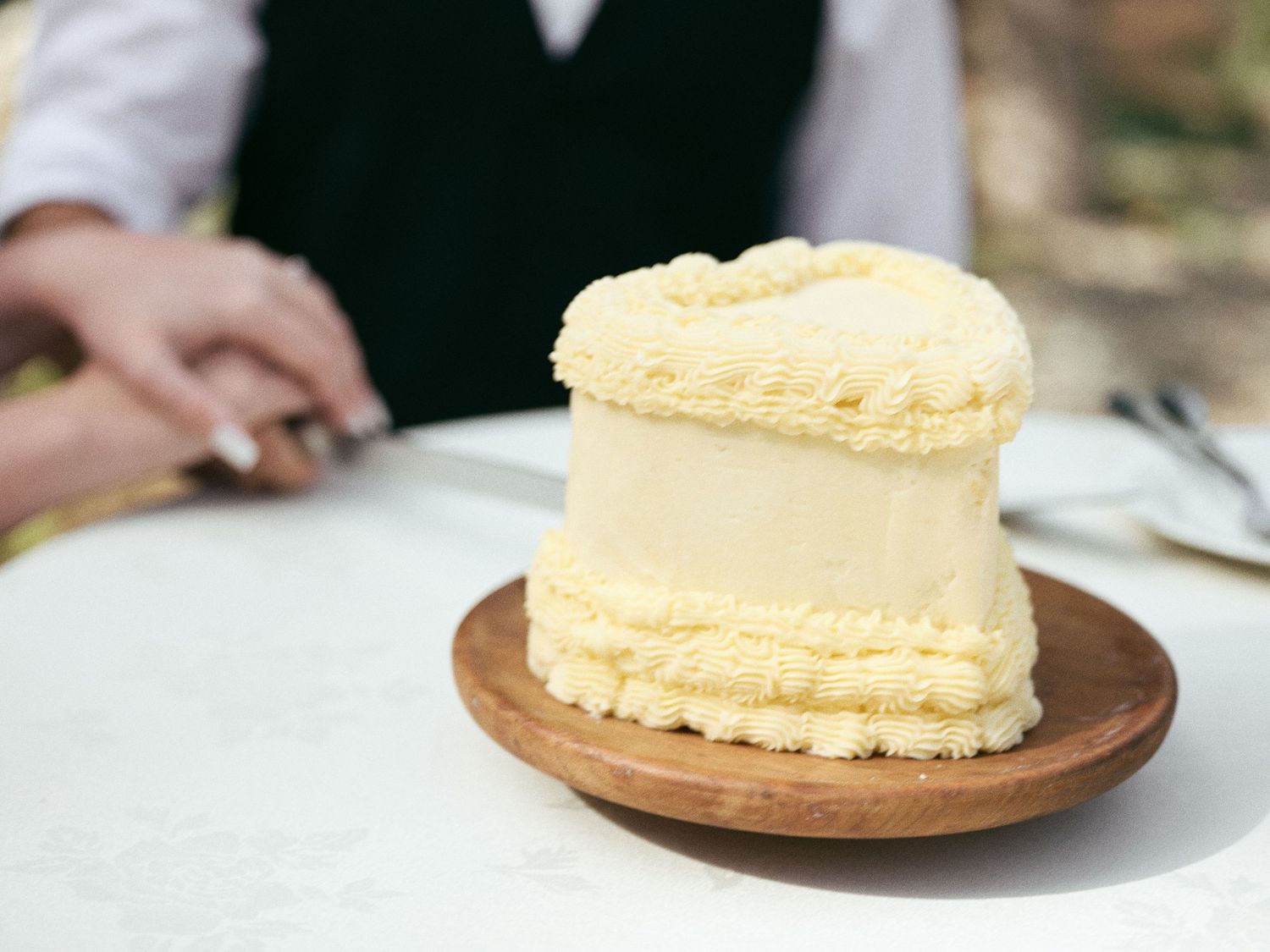 Small heart-shaped buttercream wedding cake on a rustic wooden stand set on a white outdoor table.