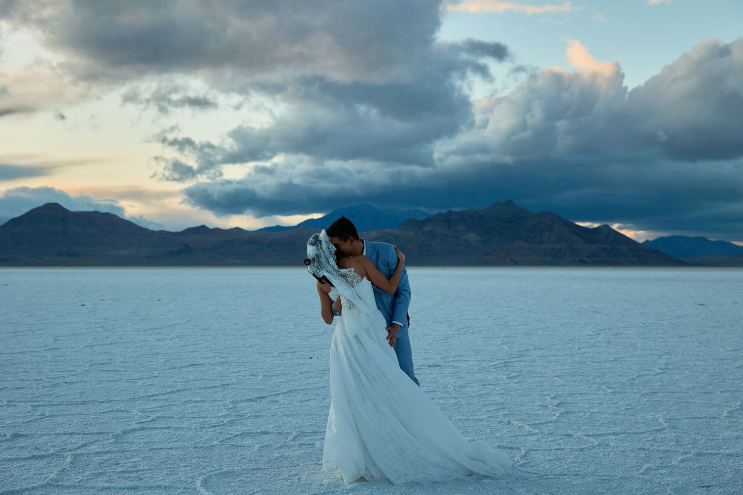 Bride and groom kiss on a vast white salt flat, her long white veil and gown flowing, with dark mountains and clouds behind.