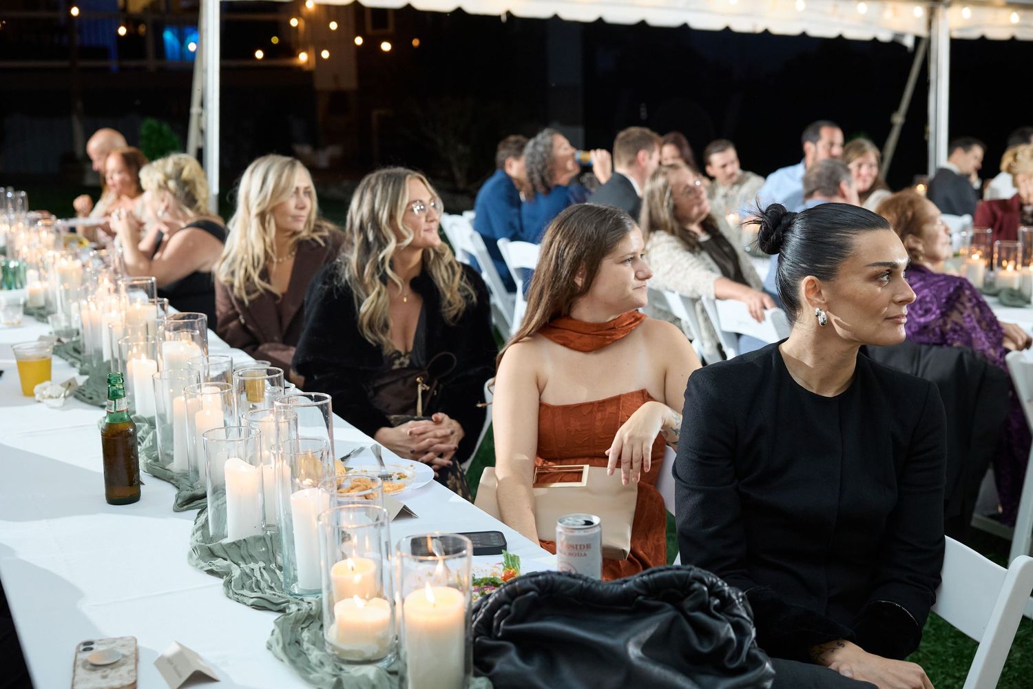 Wedding guests sit and chat around tables with glass candle holders during an evening outdoor reception under a tent.