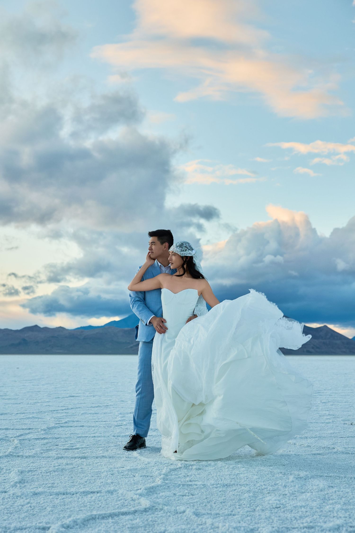Bride's white wedding gown billows in the wind as the groom in a blue suit holds her on a salt flat beneath a pastel sky.