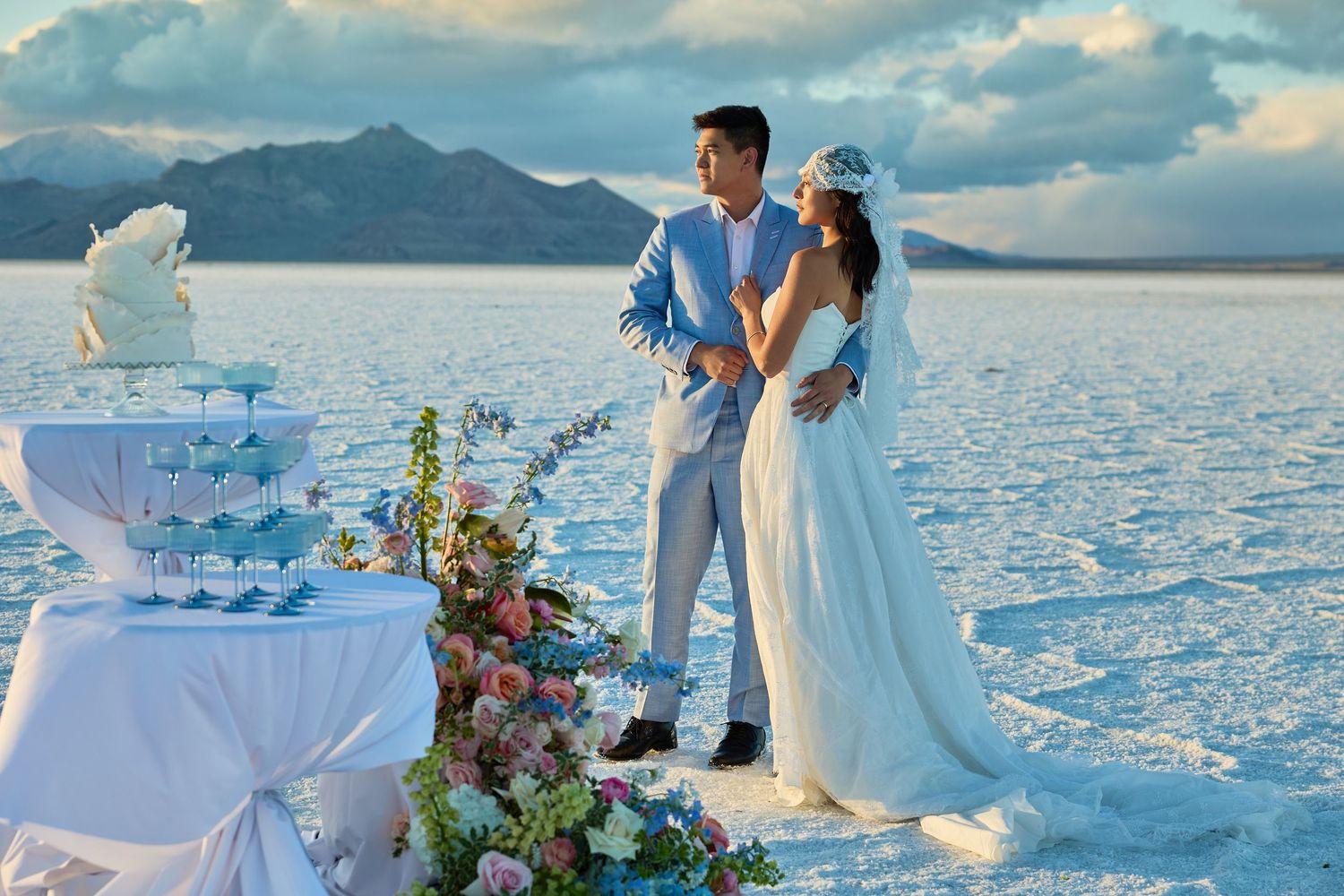 Bride and groom embrace on a white salt flat beside a floral champagne tower table under a dramatic cloudy sky.