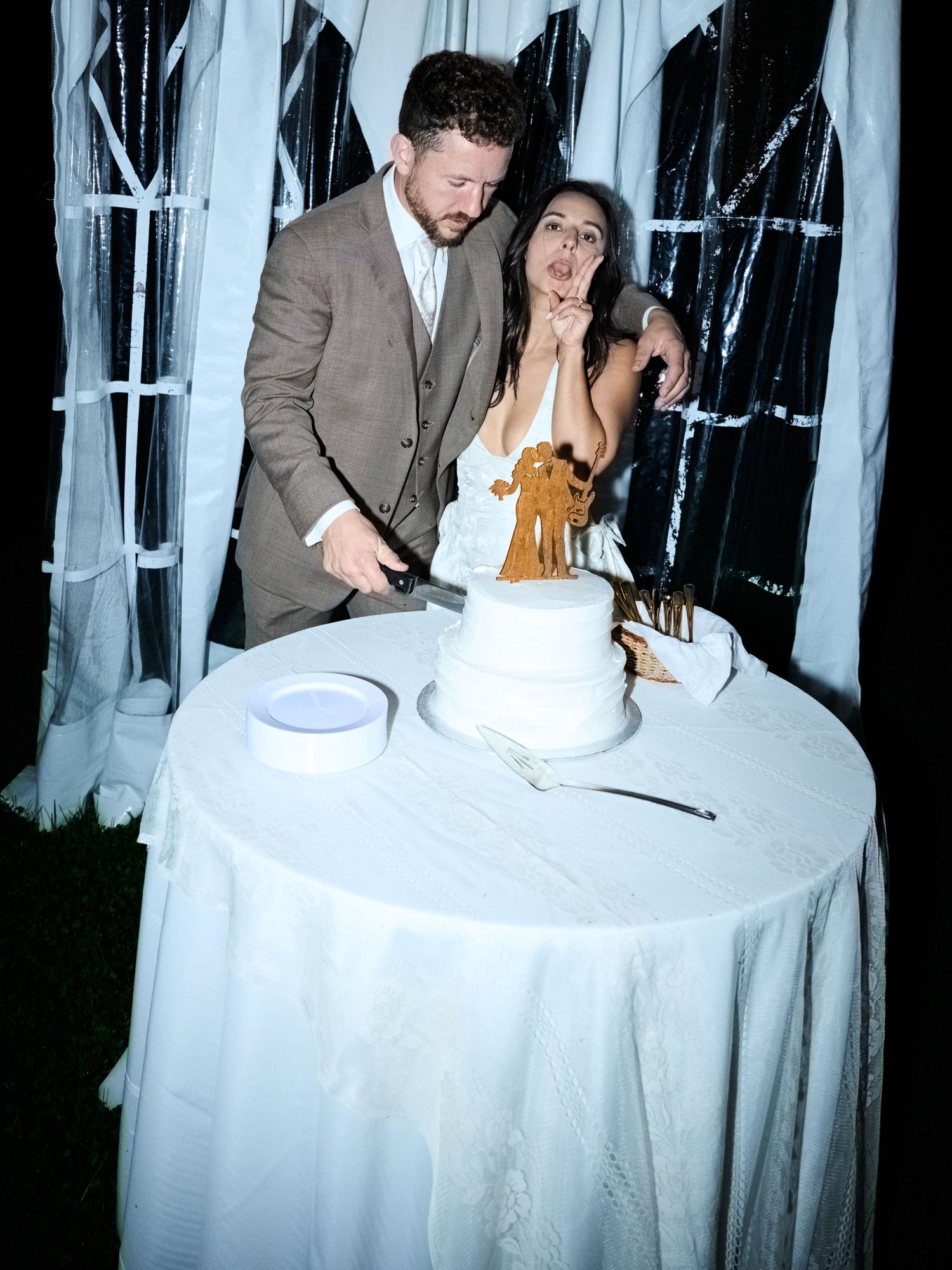 A groom in a tan suit embraces his bride in a white gown as they prepare to cut their wedding cake at a nighttime reception.