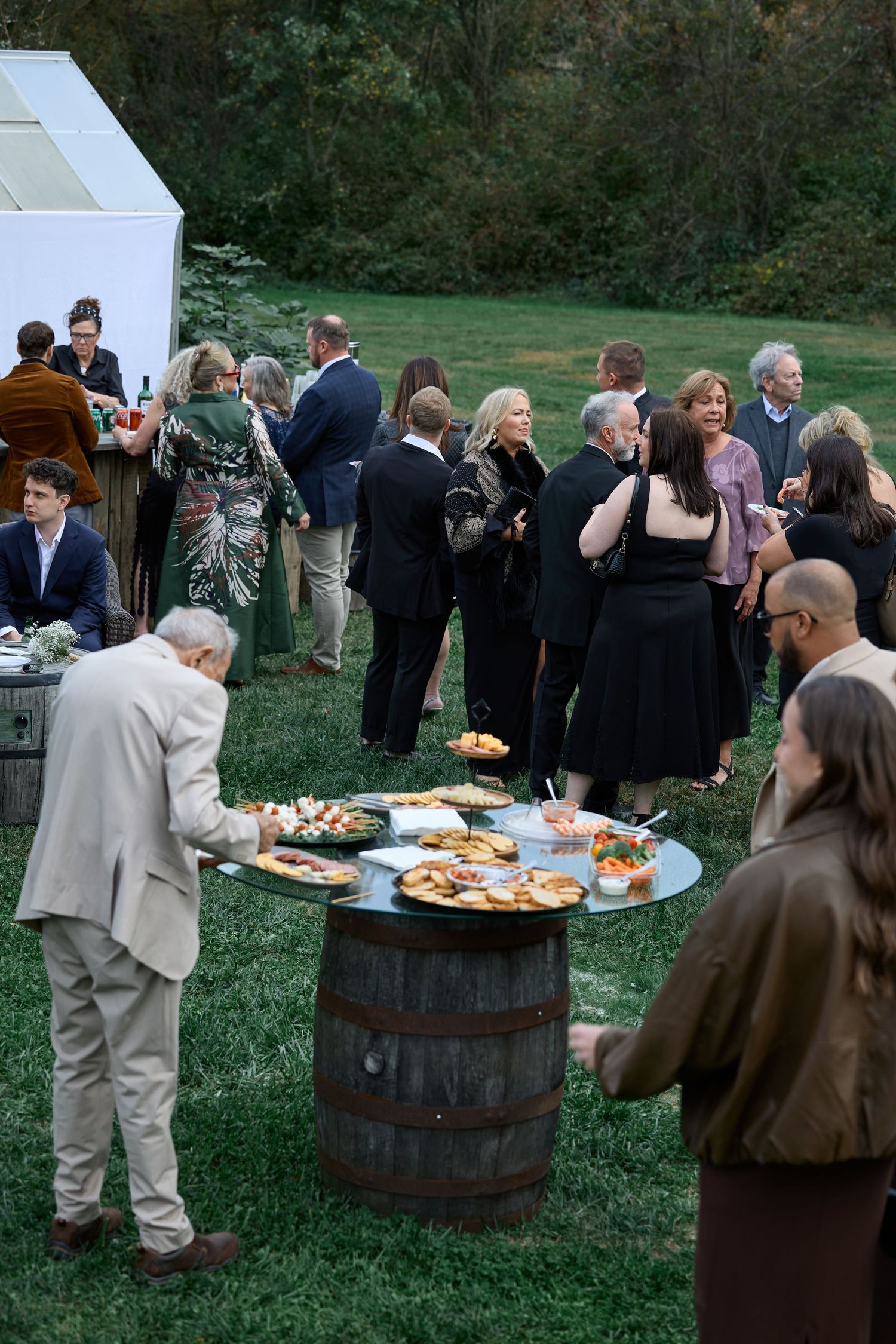 Guests gather around a wine barrel table topped with appetizer platters during an outdoor wedding cocktail hour on a green lawn.