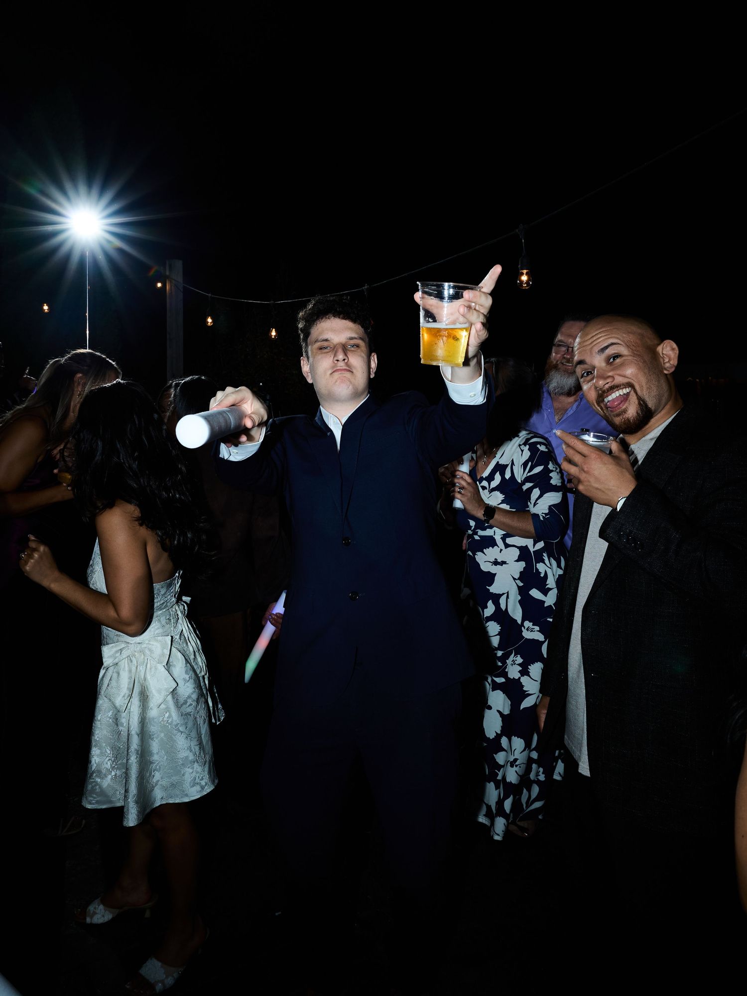 A young man in a dark suit raises a drink and glow stick while dancing with guests at a lively night wedding party.
