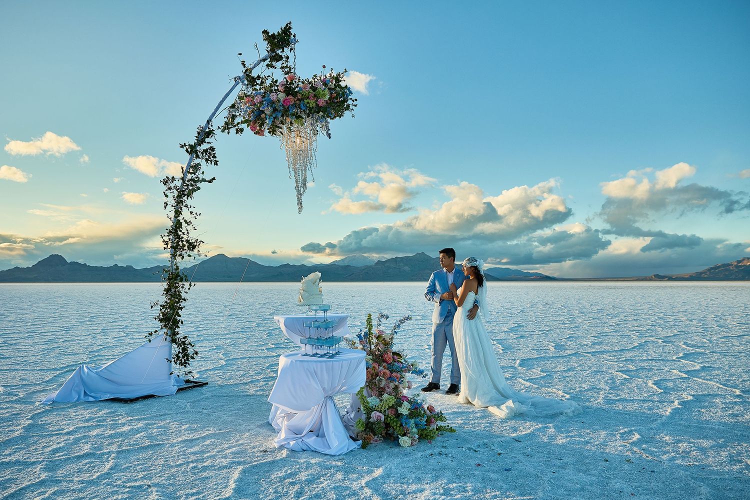 Wedding couple stands beneath a floral arch on a vast salt flat with mountains and a champagne tower in the distance.