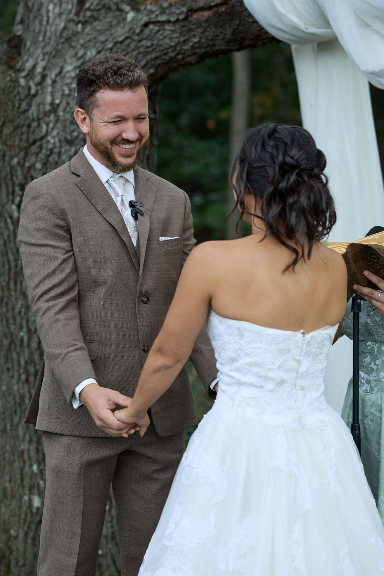 Smiling groom in a brown suit holds hands with the bride in a strapless white gown during their wedding vows.