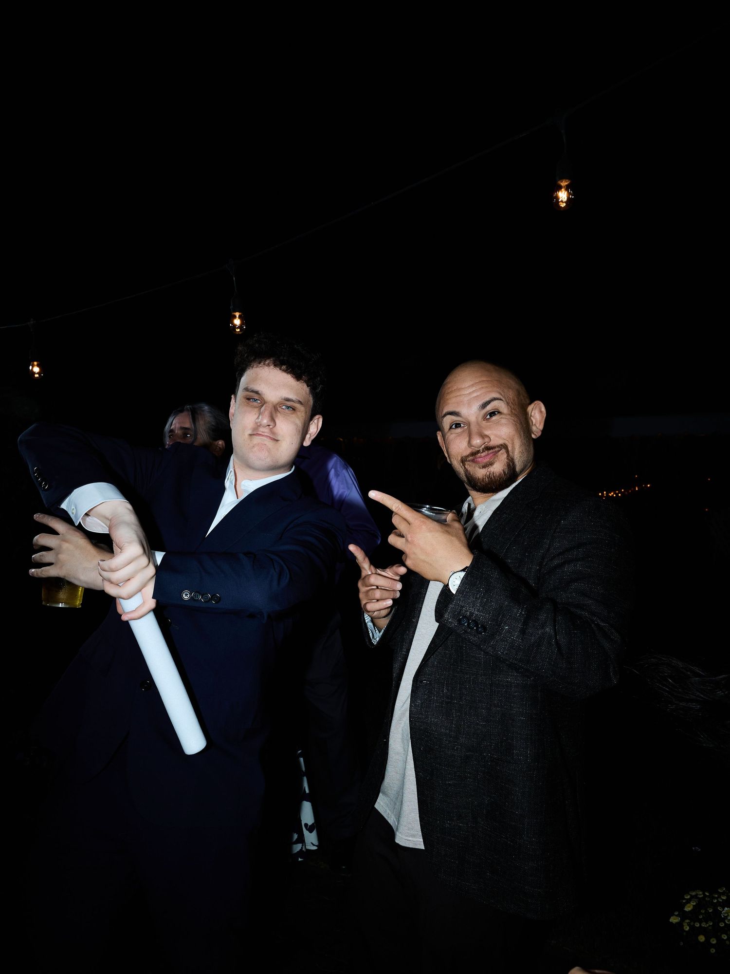 Two men in dark suits pose playfully at a nighttime wedding reception, one pointing at the camera.