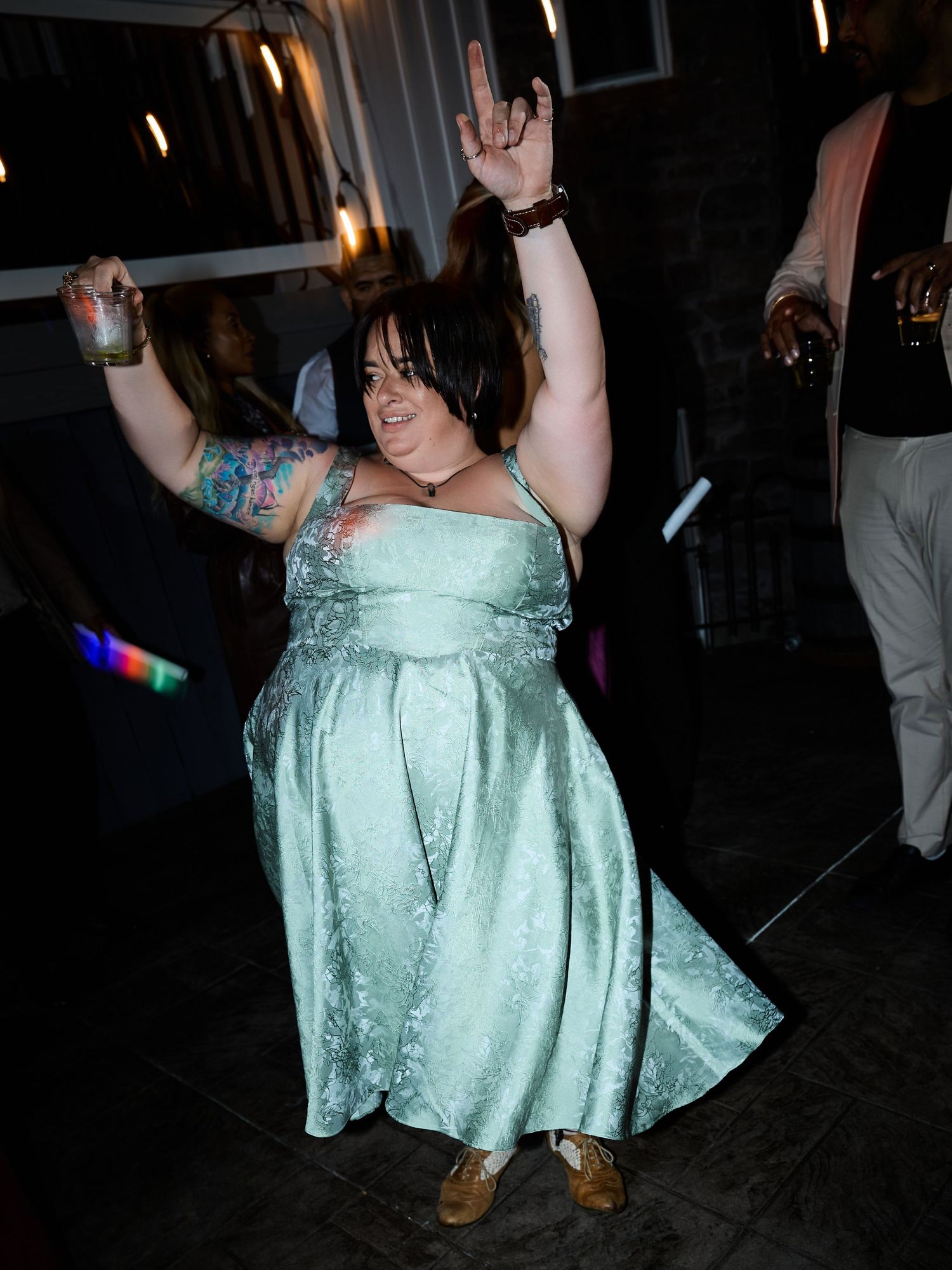A woman in a mint green satin dress with arm tattoos dances joyfully, holding a drink and making a rock hand sign at a wedding.