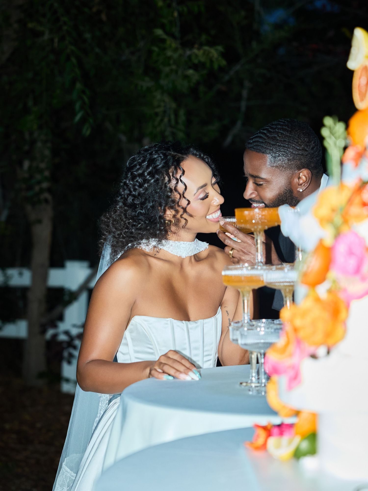 A joyful bride and groom sip champagne from coupe glasses next to a floral wedding cake decorated with orange and pink blooms at night.