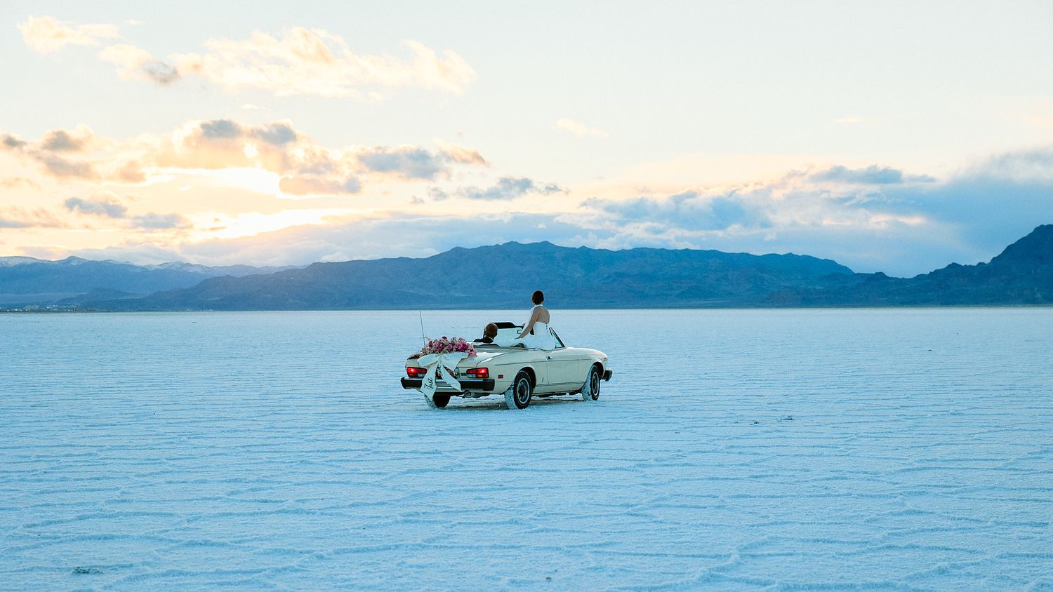 A lone person stands in a vintage convertible with floral decor on a vast pale salt flat at sunset with distant mountains.