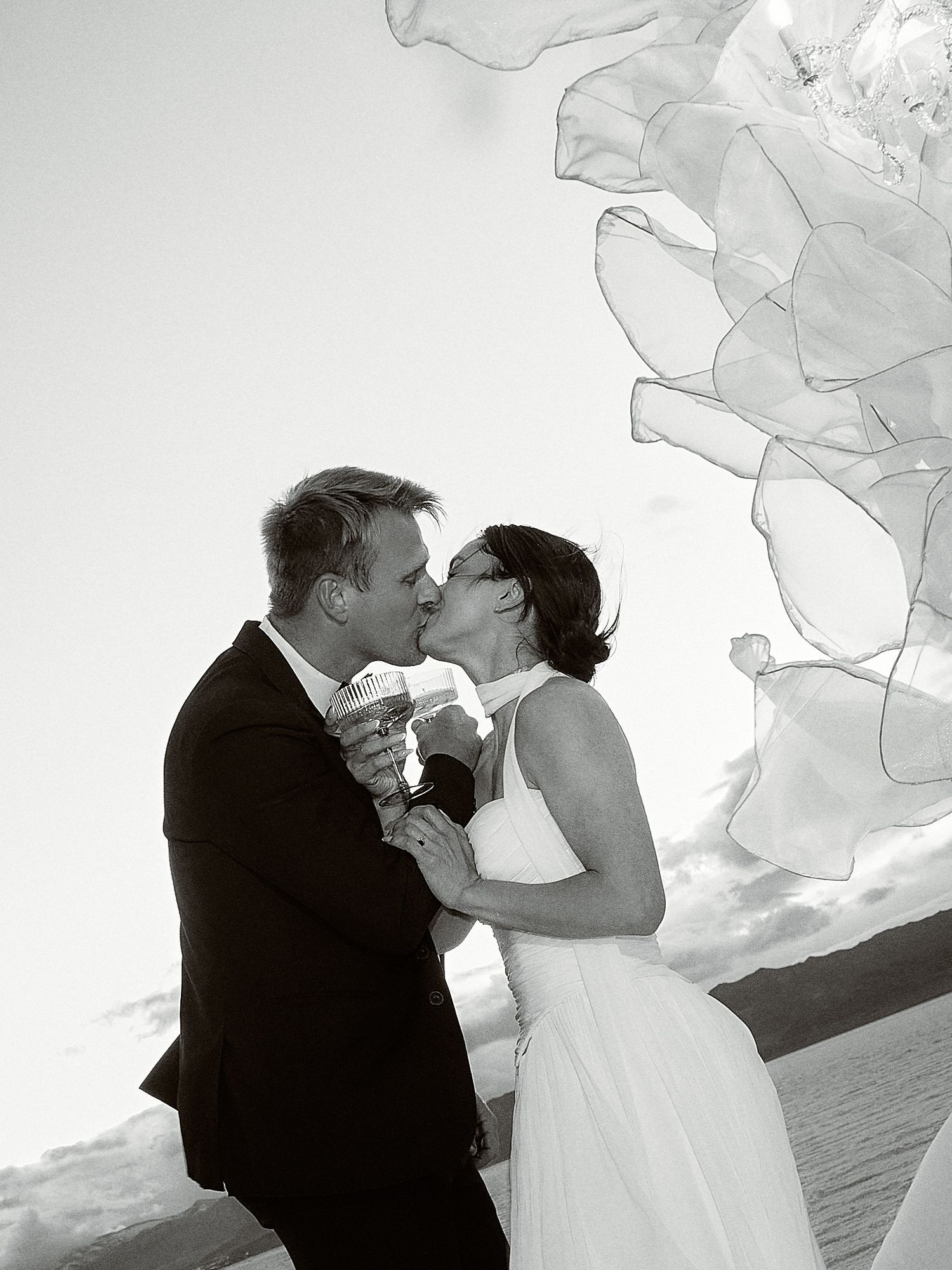 Black and white photo of a newlywed couple kissing while holding a glass, with flowing sheer fabric decor framing the sky.