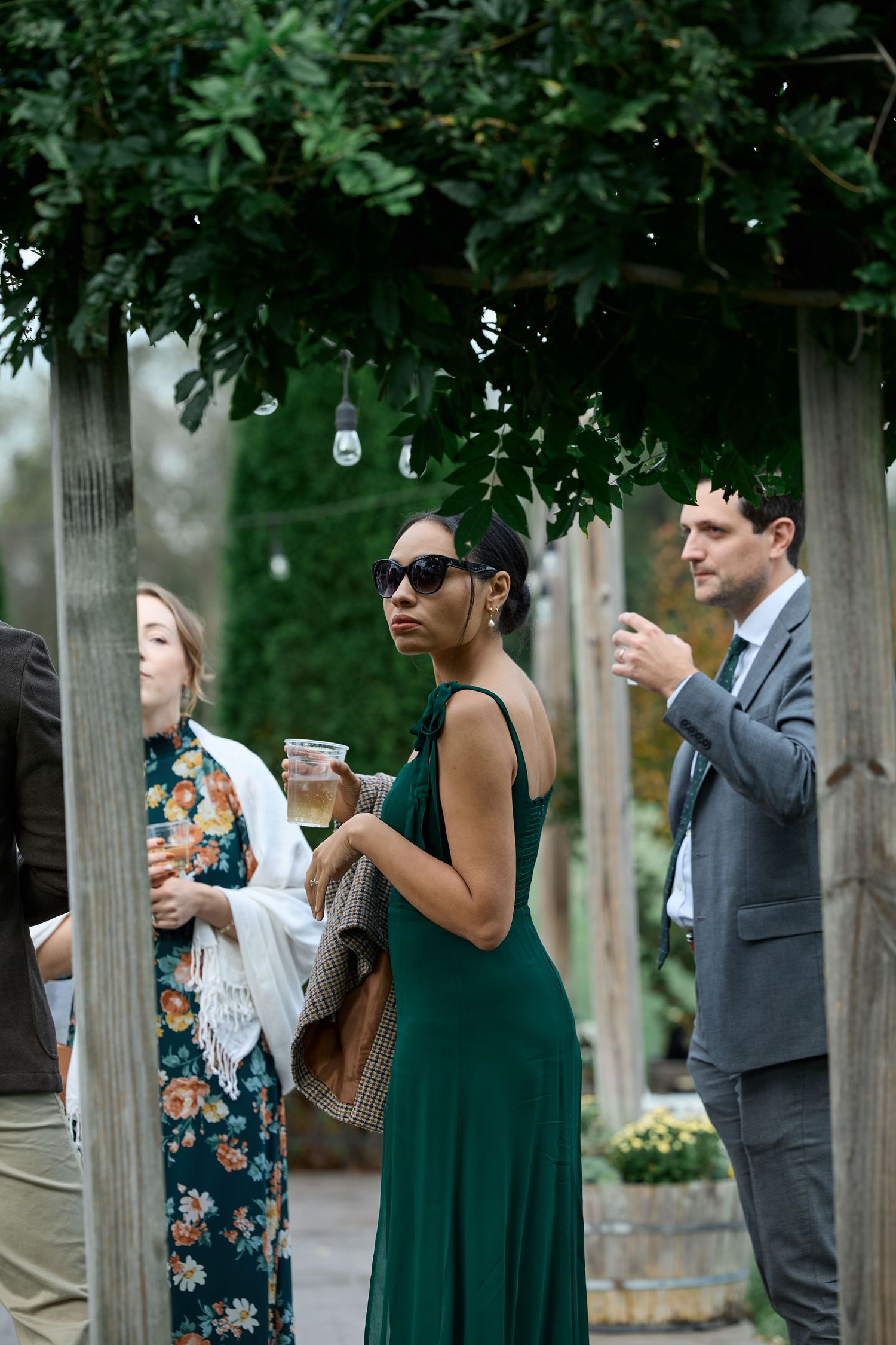 A woman in a green dress and sunglasses holds a drink beneath a leafy pergola while guests chat nearby at an outdoor event.