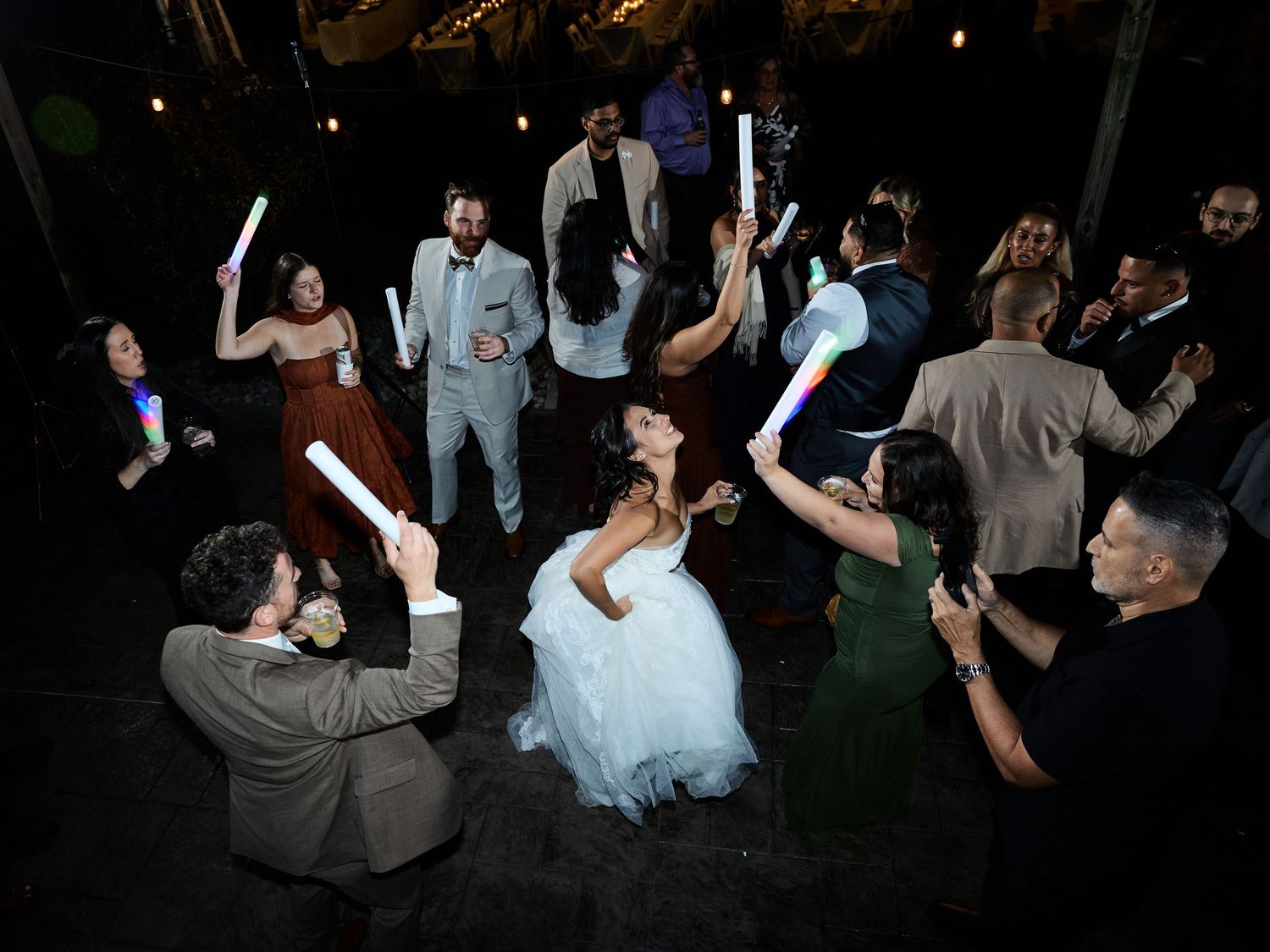 Overhead view of a bride in a white gown dancing in a crowd of wedding guests waving glowing light sticks at night.
