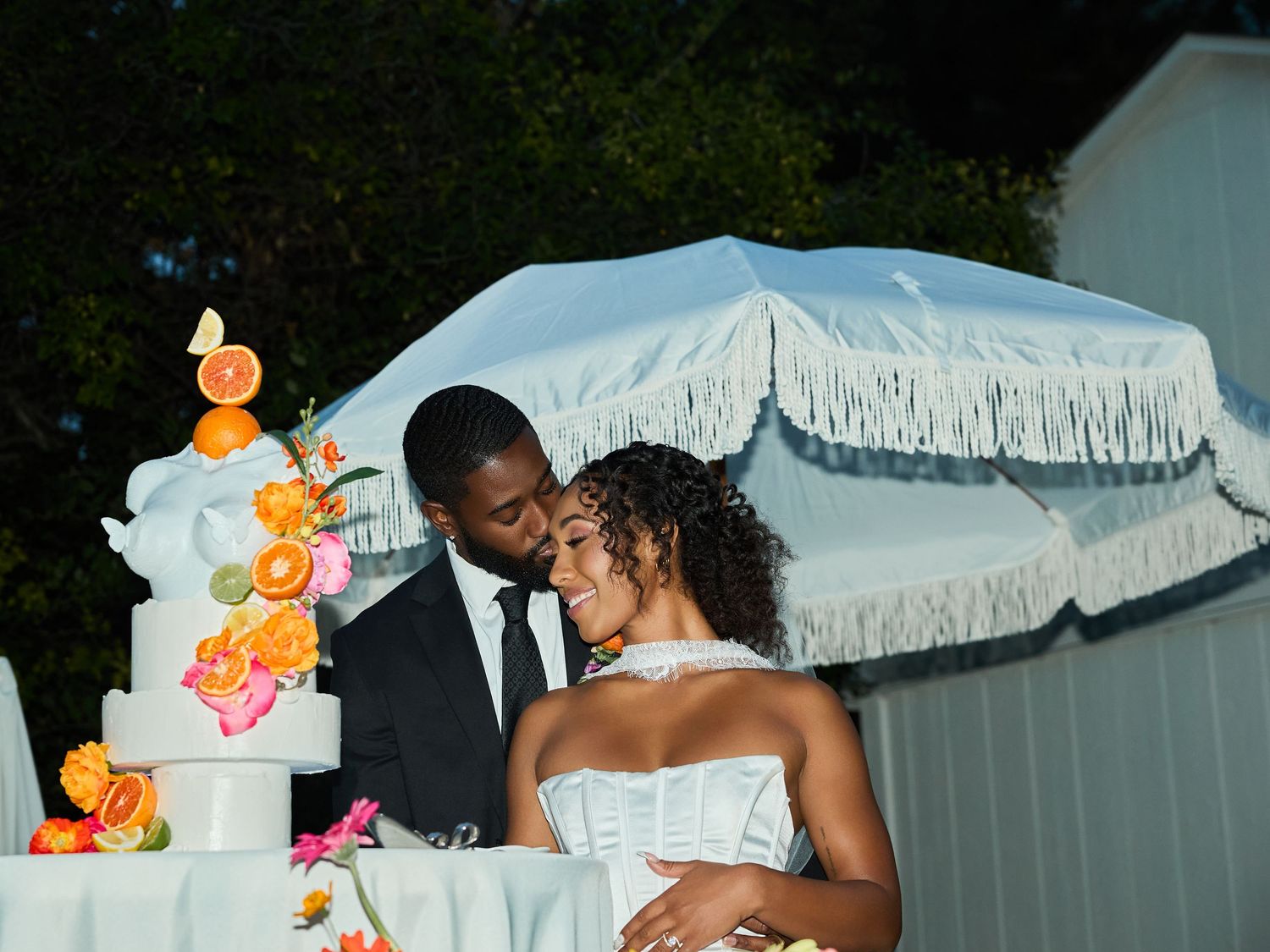 Groom kisses his bride's cheek under a white fringed umbrella next to a wedding cake adorned with oranges and pink flowers.